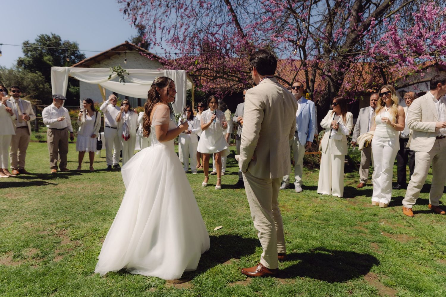 Bride and groom facing each other during an outdoor wedding ceremony with guests gathered around.