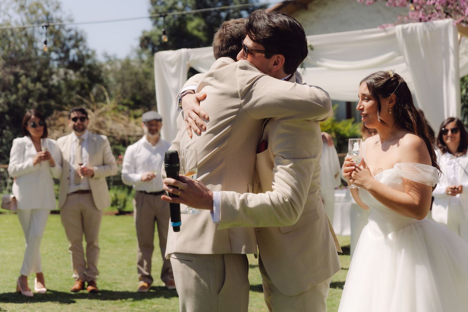Groom embracing a guest while bride smiles holding champagne at an outdoor wedding reception.