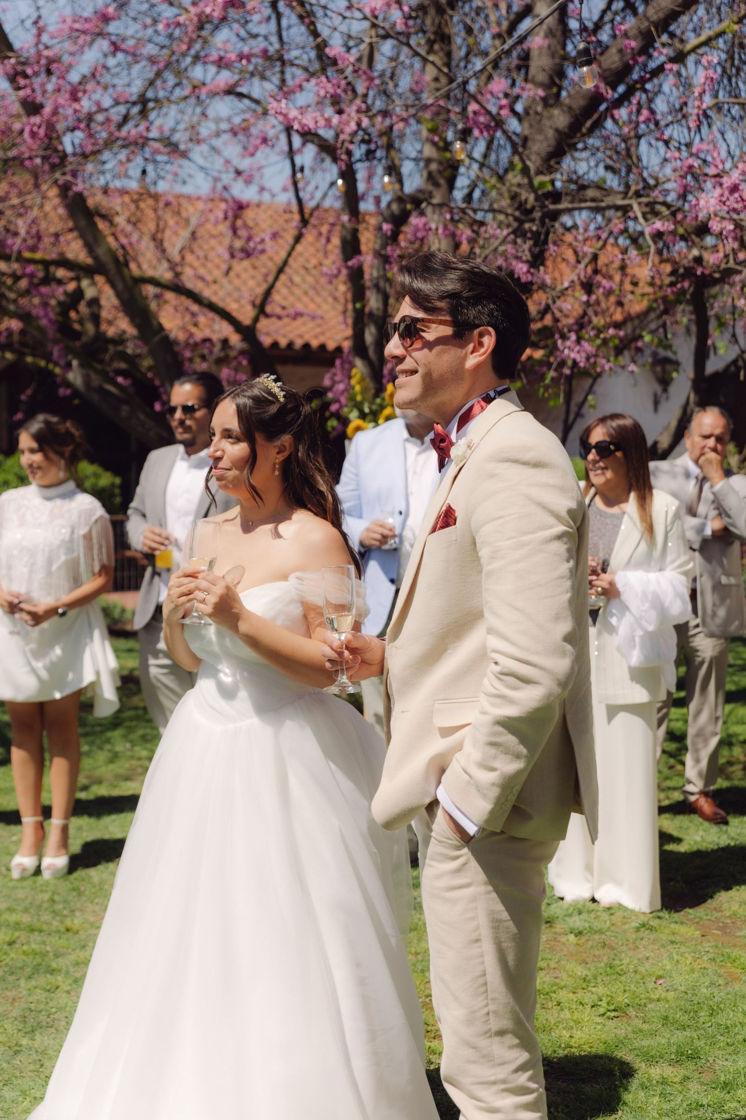 Bride and groom holding champagne glasses at an outdoor wedding with pink blossoming trees.