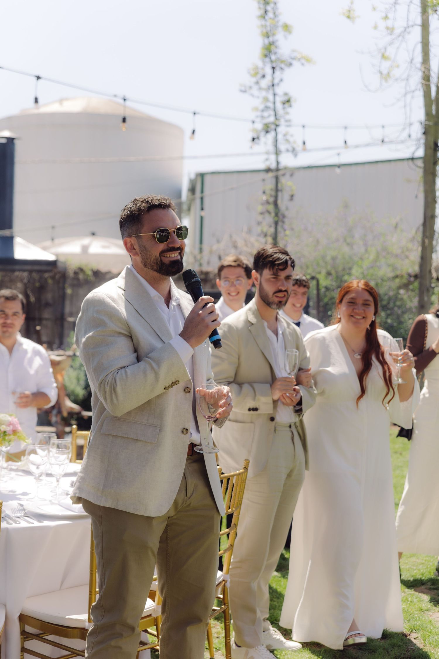 Man giving a speech at an outdoor wedding reception with guests standing nearby.