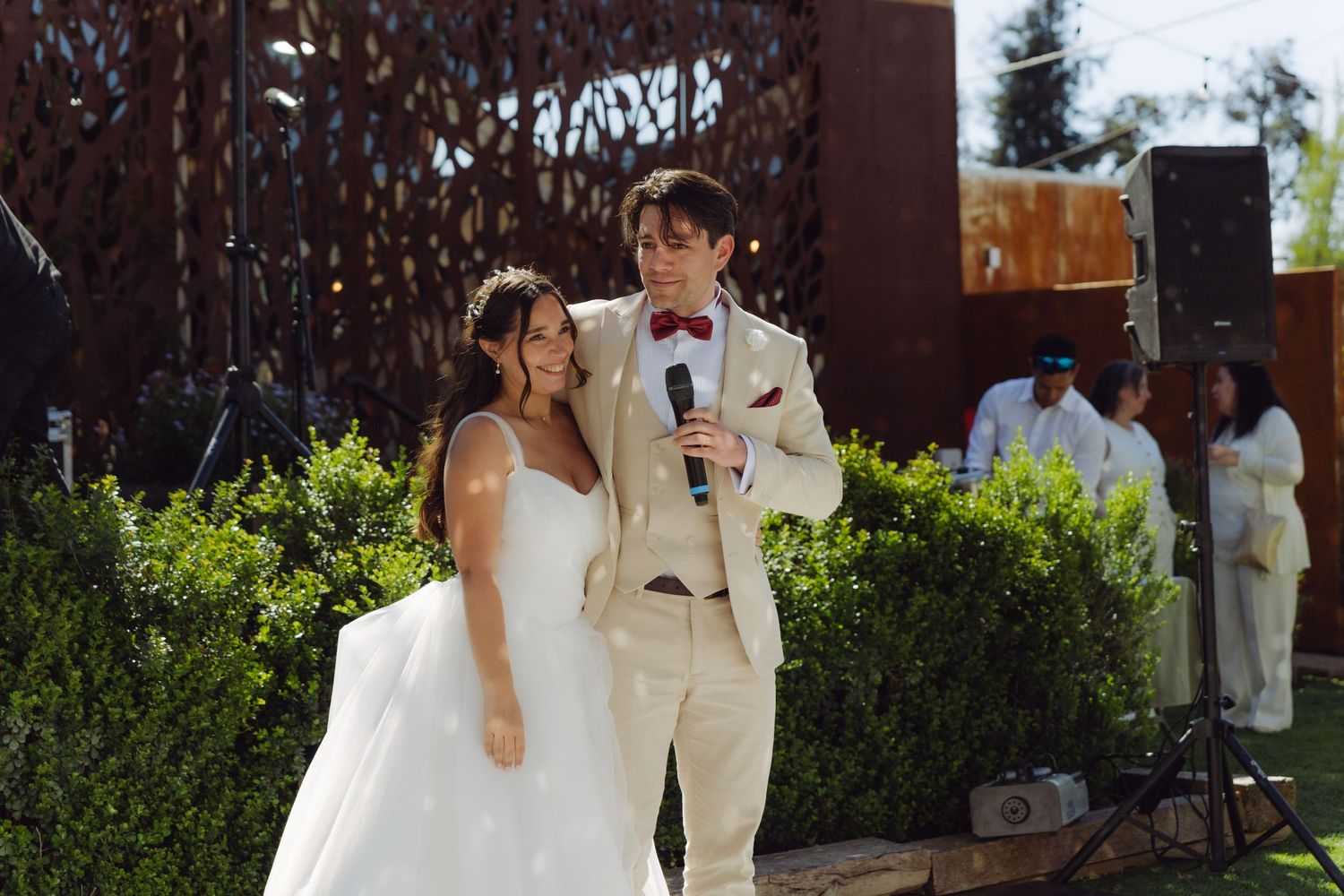 Bride and groom smiling while groom speaks into a microphone at their outdoor wedding reception.