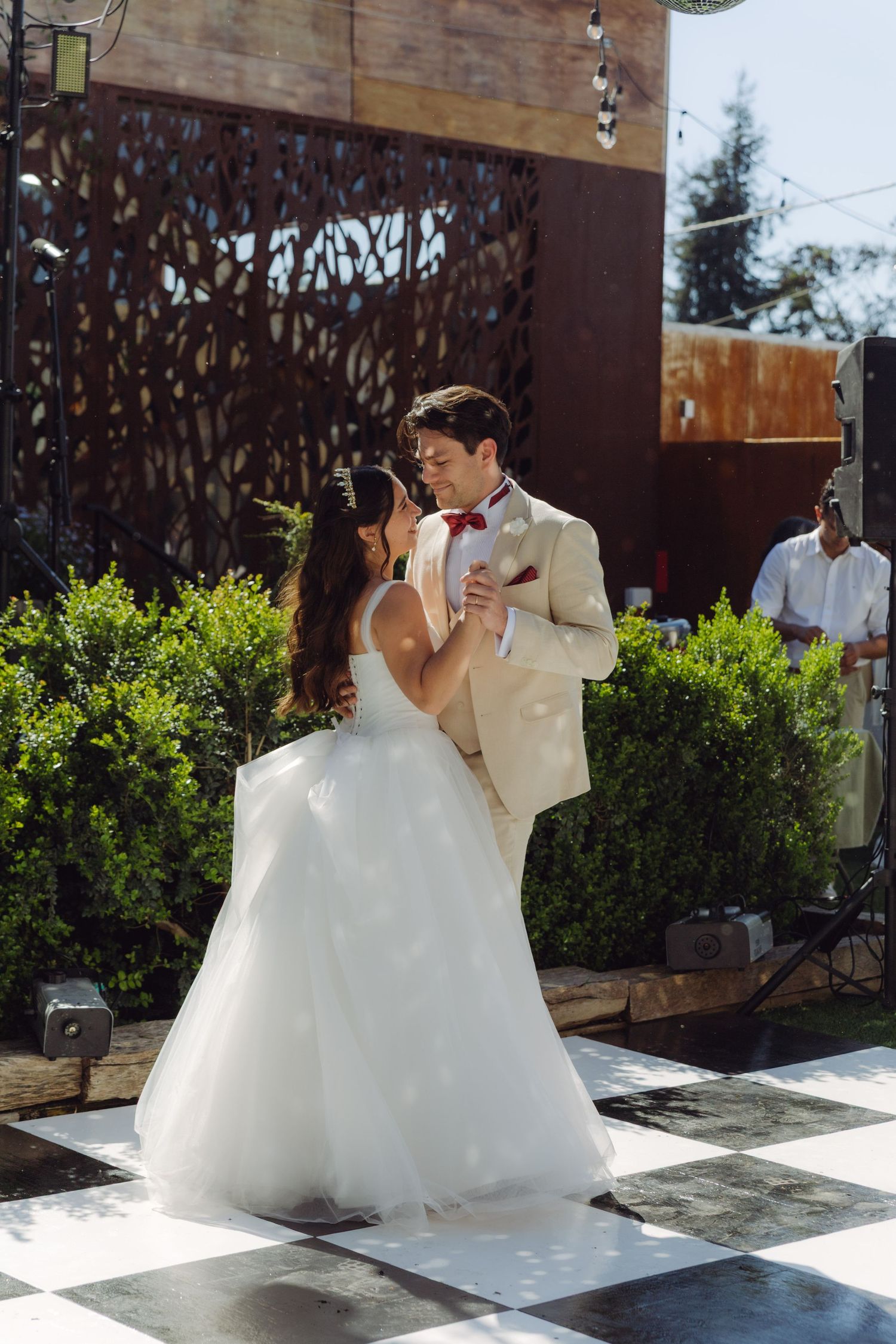 Bride and groom share their first dance outdoors on a checkered dance floor surrounded by lush greenery.
