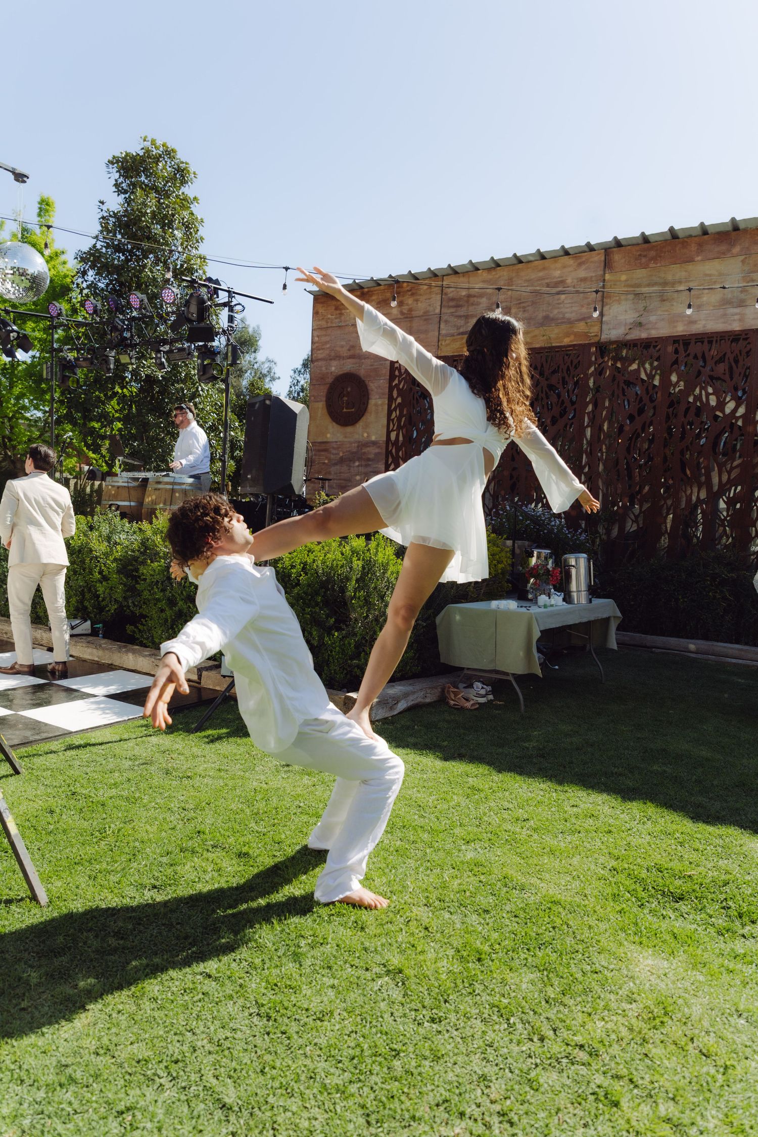 Two performers in white execute an acrobatic dance lift on a green lawn during an outdoor wedding reception.