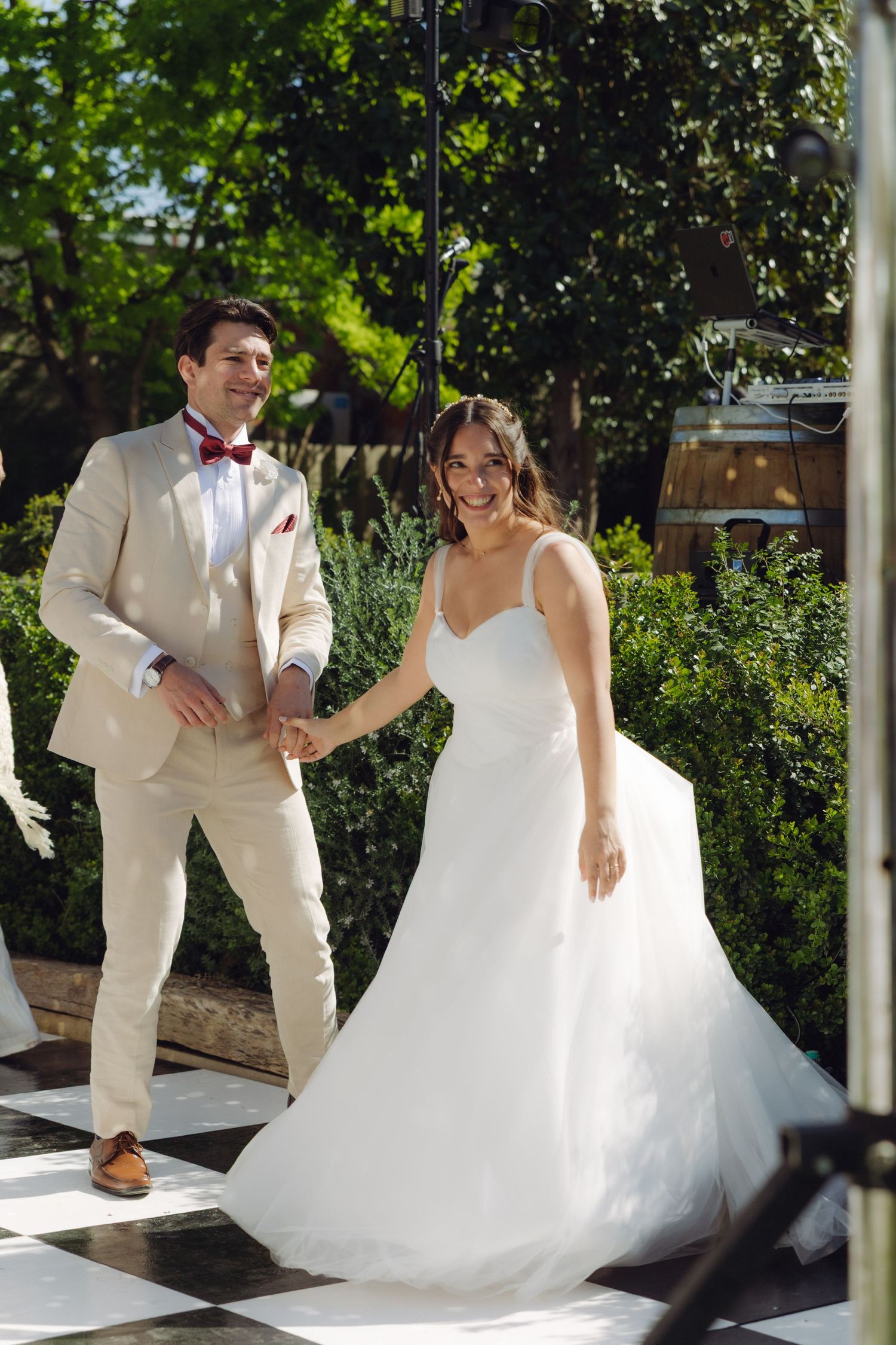 Smiling bride in white gown and groom in beige suit hold hands on a checkered outdoor dance floor.
