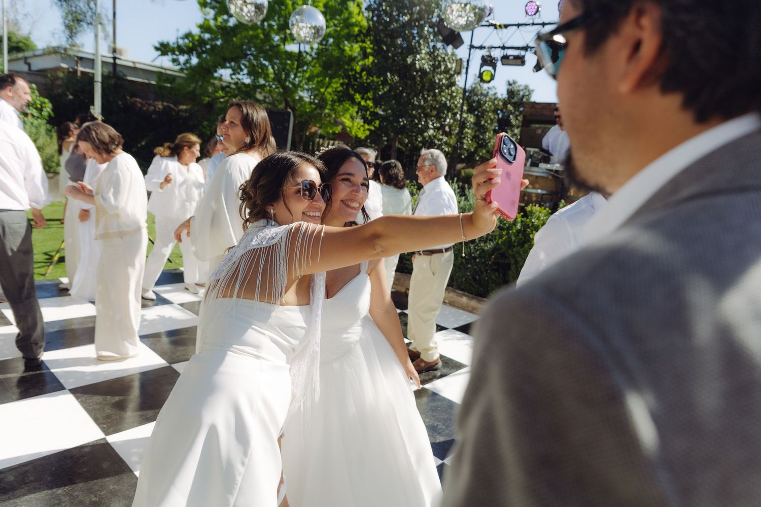 Bride in white wedding gown takes a selfie with a guest on the dance floor at an outdoor reception.