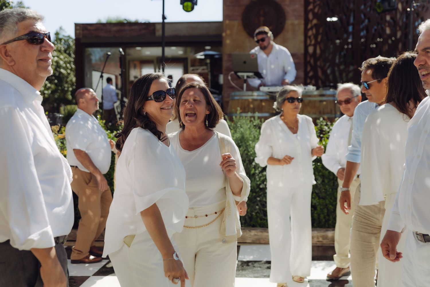 Wedding guests in white attire laugh and celebrate together outdoors at a sunny daytime reception.