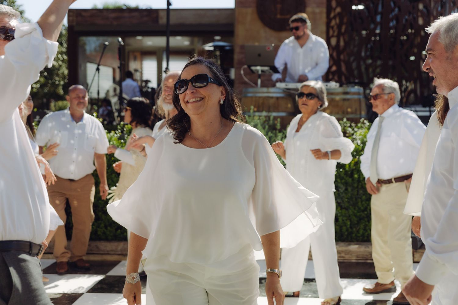 Joyful woman in white outfit smiles and dances on a checkered floor surrounded by wedding guests.