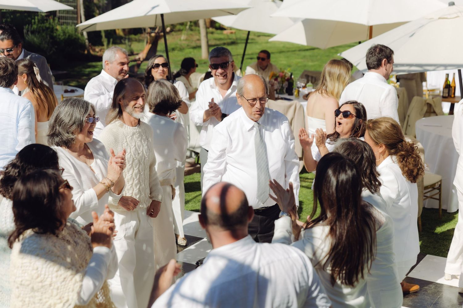 Wedding guests in white attire mingle and celebrate outdoors under large umbrellas at a sunny reception.