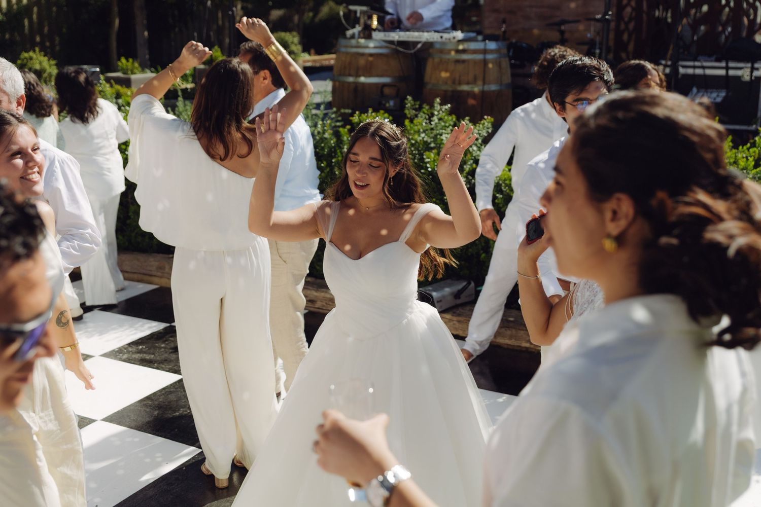 Bride in white gown dancing joyfully with guests at outdoor wedding reception on checkered dance floor.