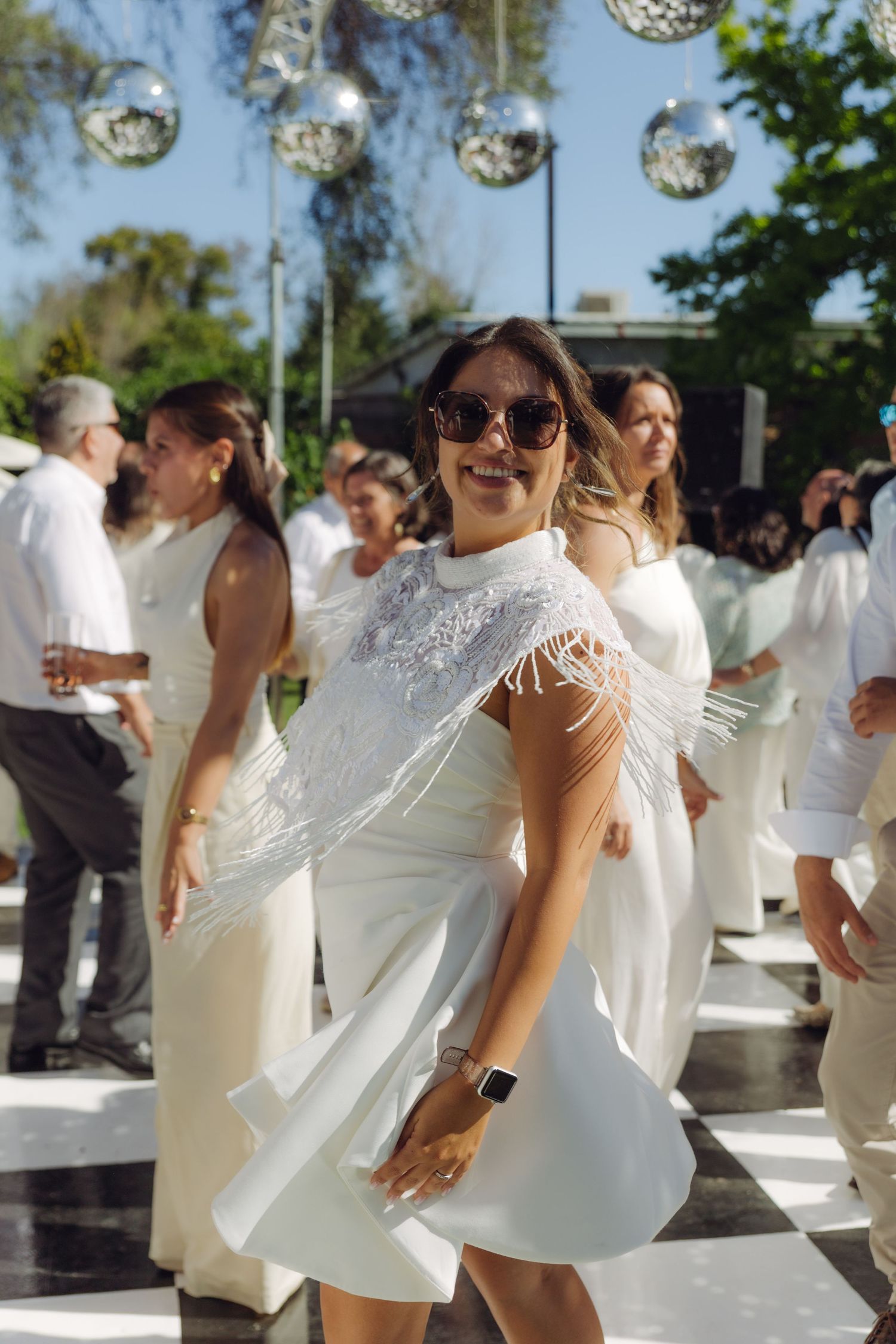 Woman in white fringed dress dancing at outdoor wedding party under hanging disco balls.