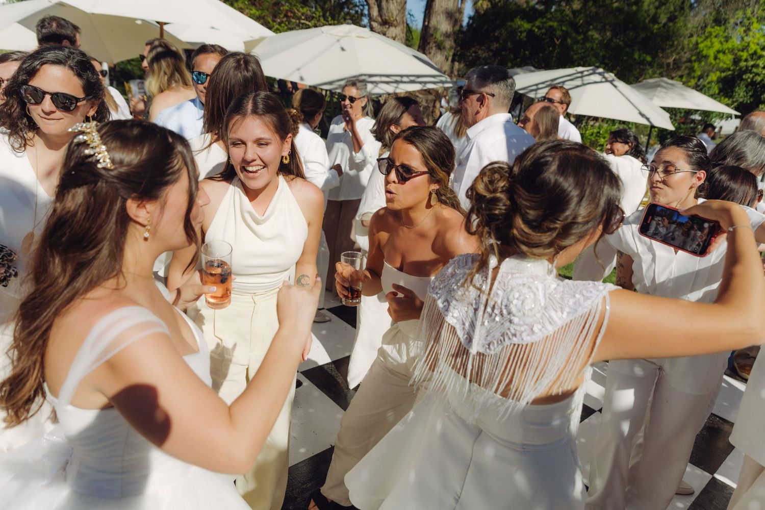 Wedding guests in all-white attire dancing and taking selfies at outdoor daytime celebration.