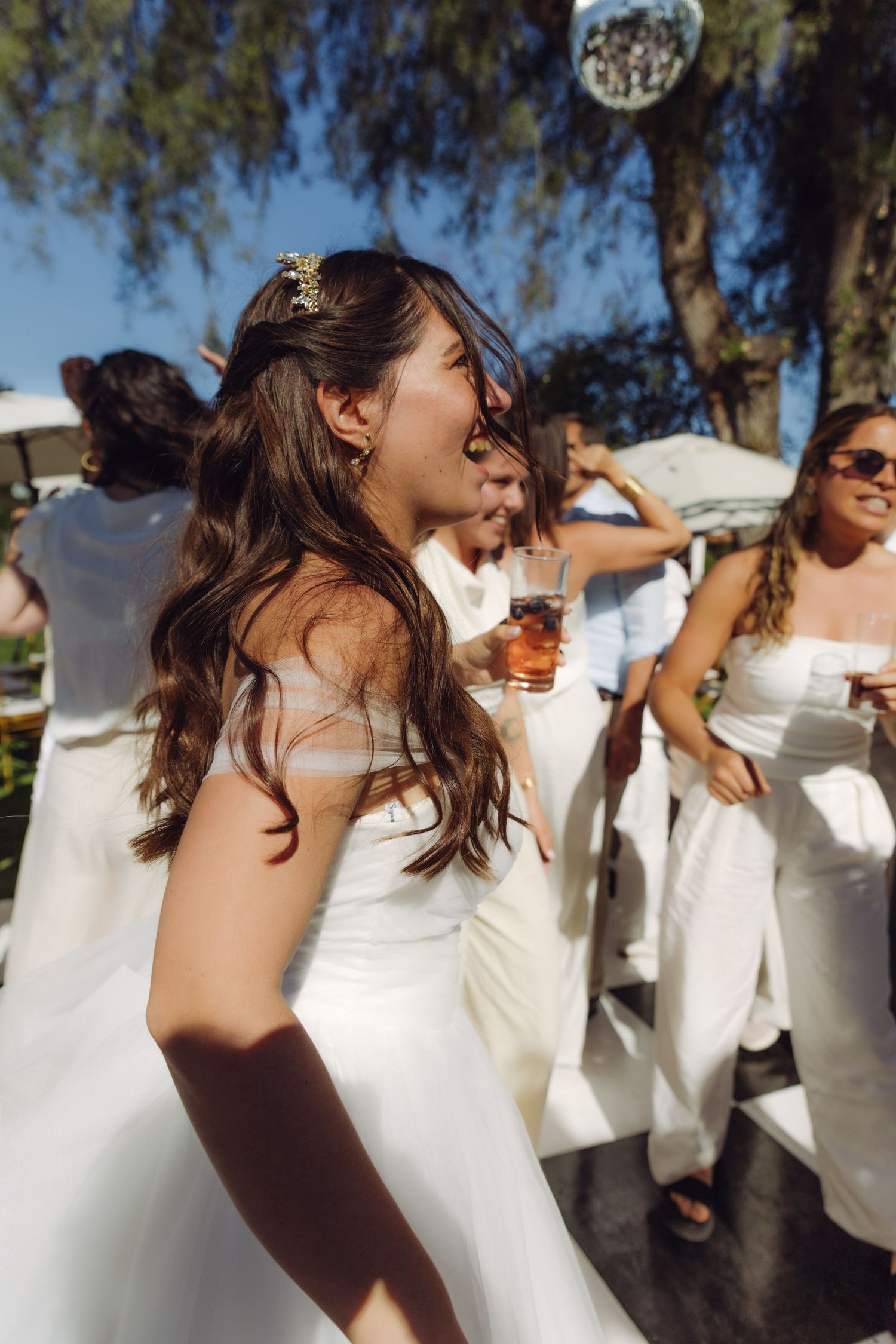 Bride with hair accessory dancing joyfully holding drink at outdoor wedding reception.