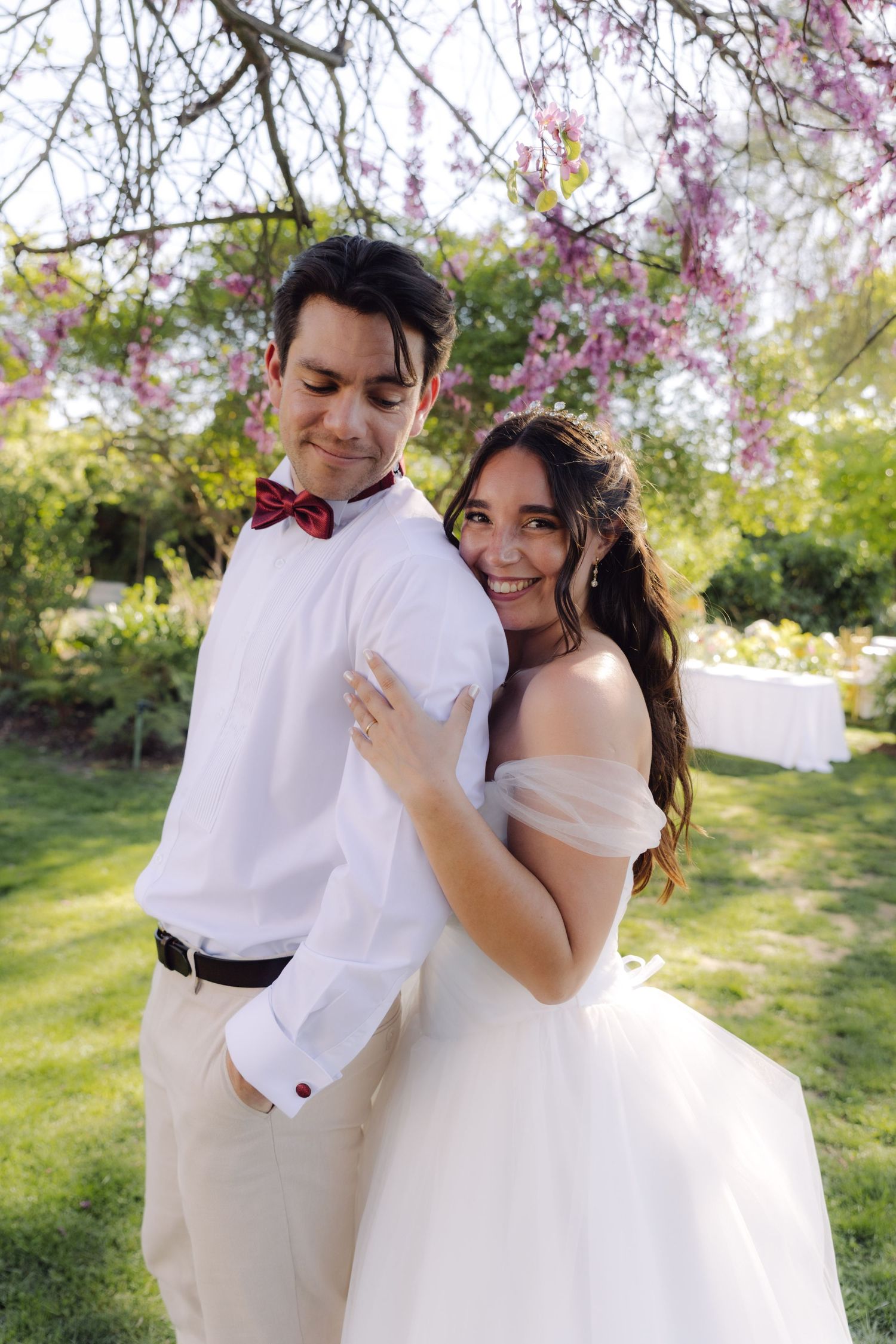 Smiling bride hugging groom from behind under flowering cherry blossom trees during outdoor wedding celebration.