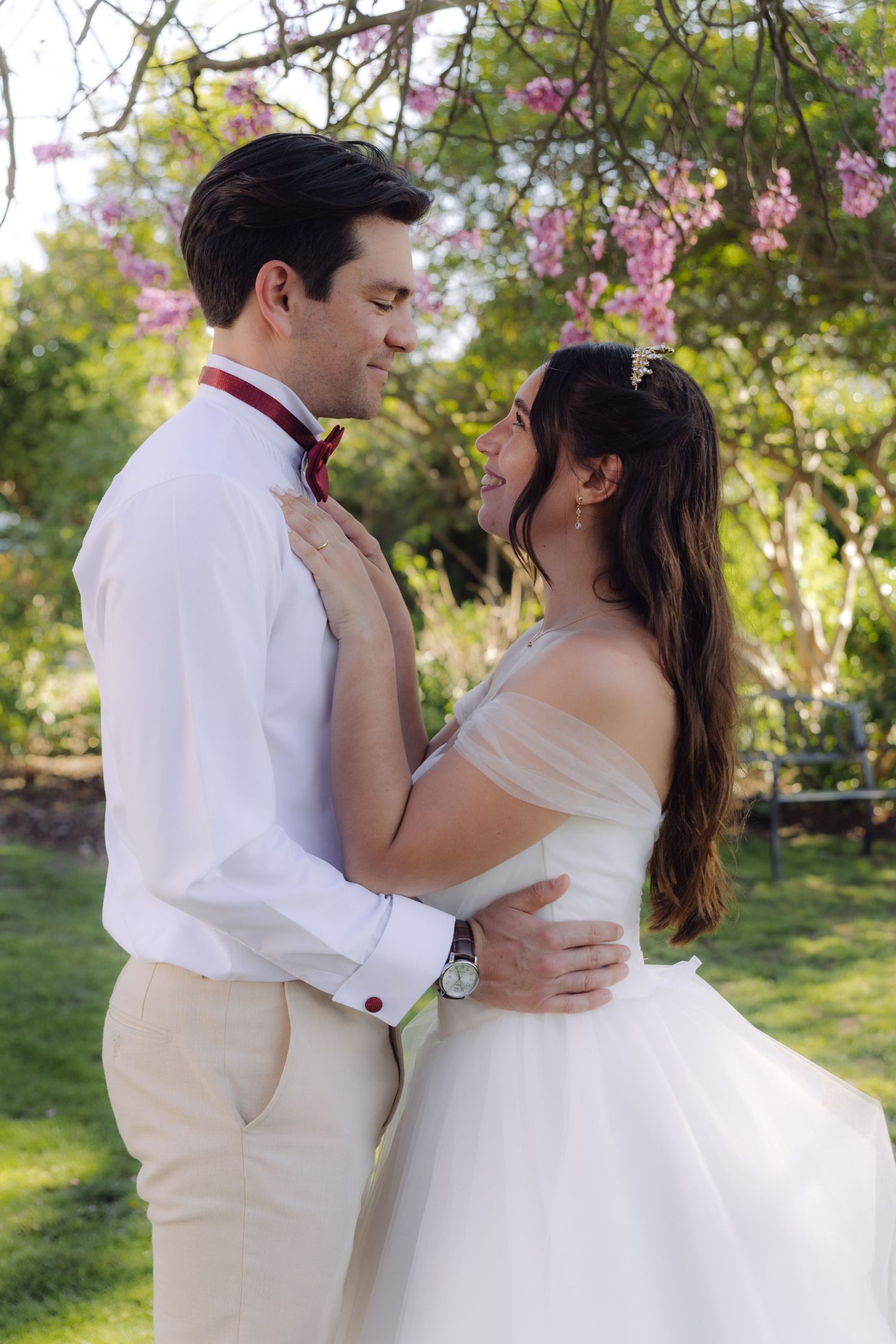 Bride and groom embracing tenderly under blooming pink spring trees in a sunlit garden wedding setting.