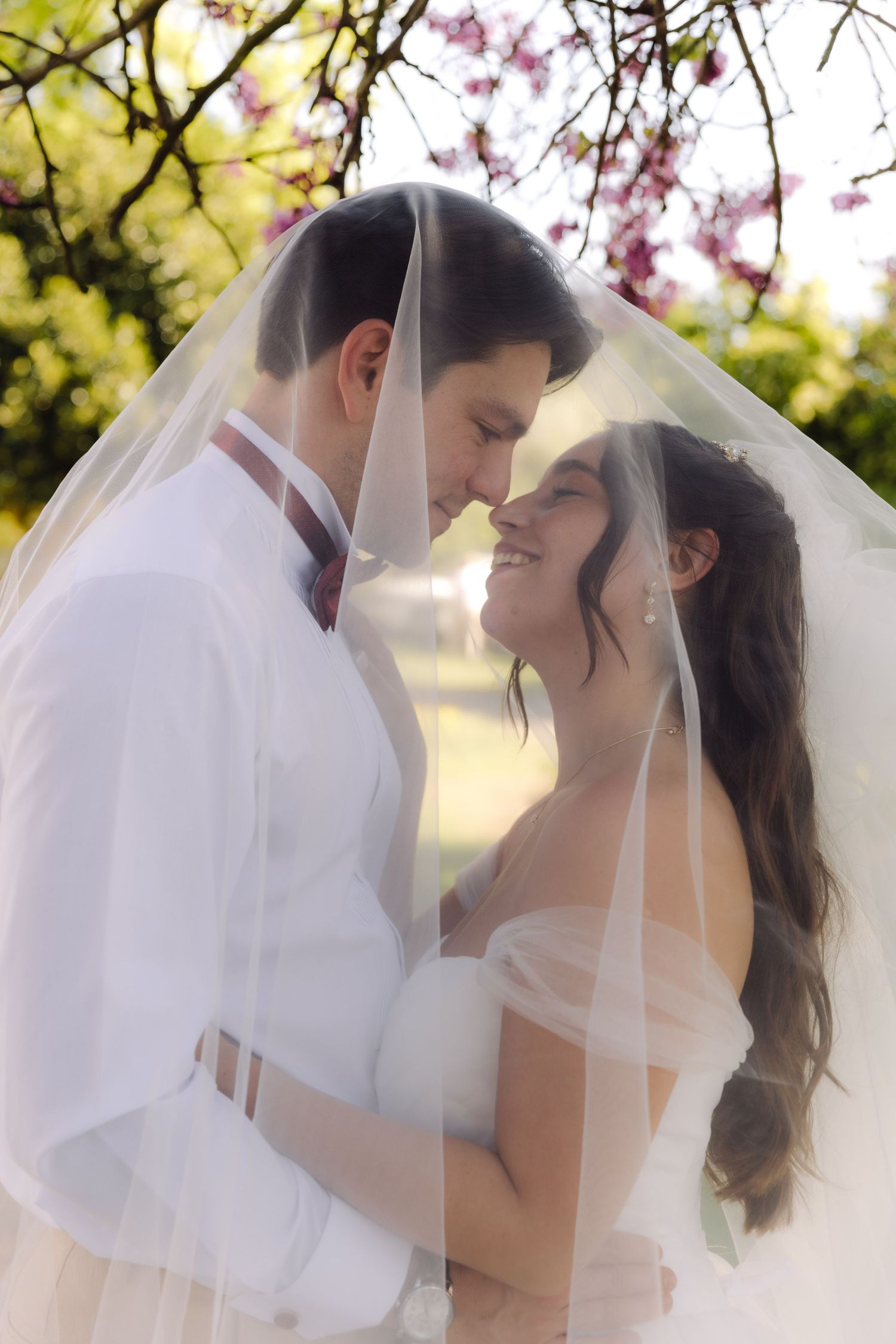 Bride and groom share romantic moment under white wedding veil surrounded by blooming spring trees.