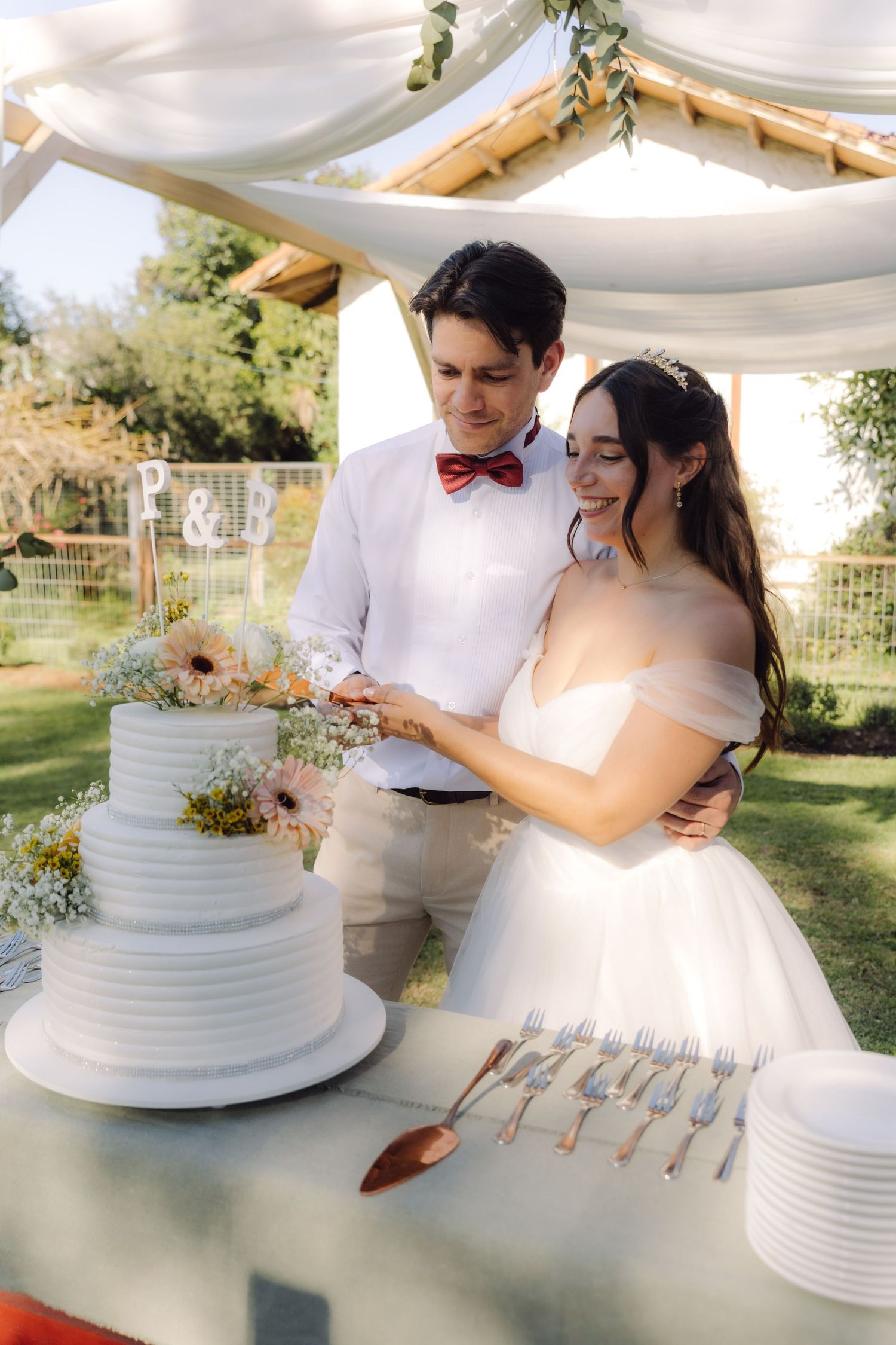 Bride and groom cutting their three-tier white wedding cake outdoors under a draped canopy.