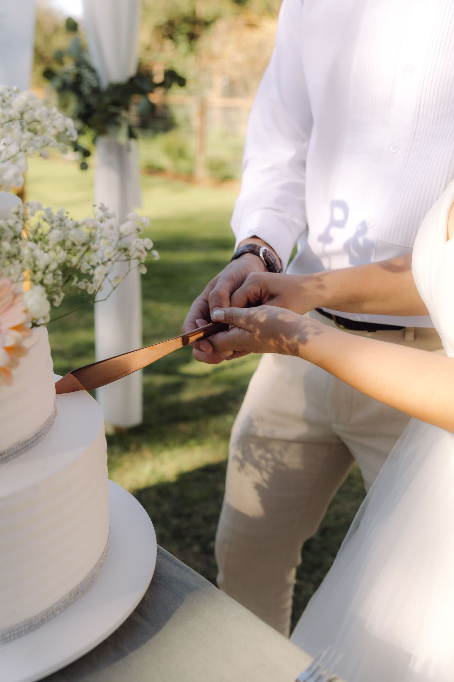 Close-up of bride and groom hands cutting white wedding cake with a gold knife outdoors.
