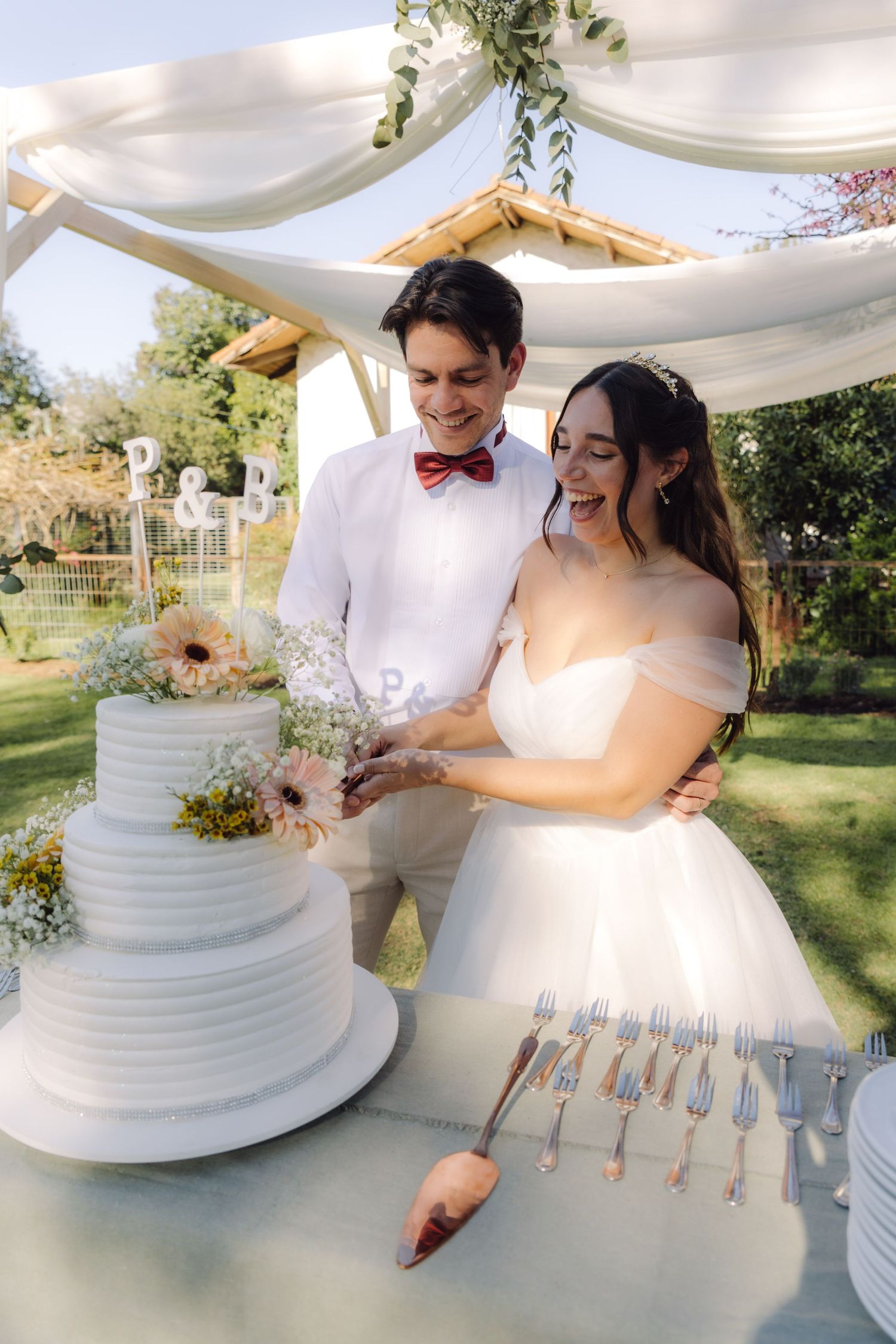 Smiling bride and groom cutting their elegant three-tier white wedding cake at outdoor reception.