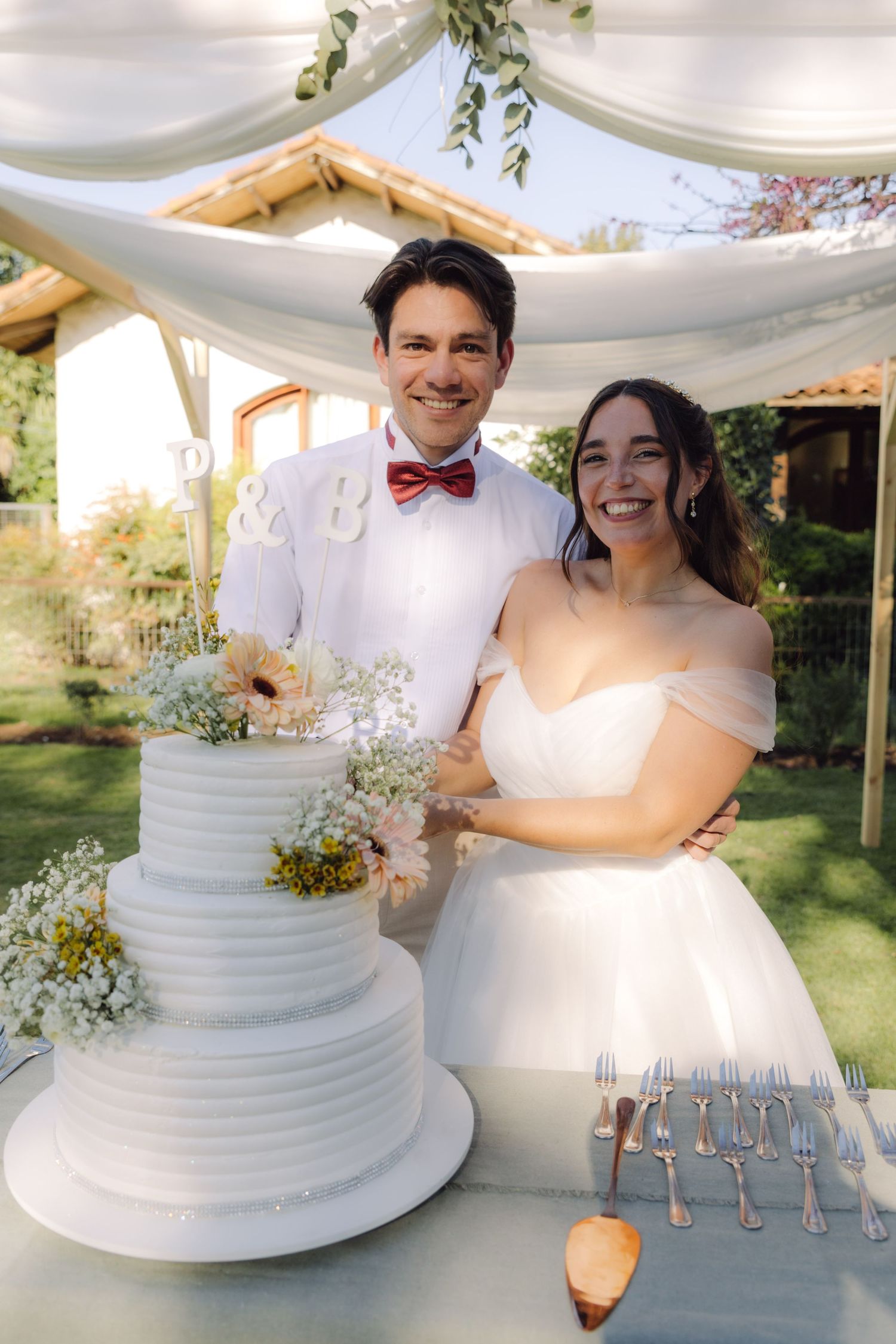 Happy bride and groom posing together beside their three-tier white wedding cake outdoors.
