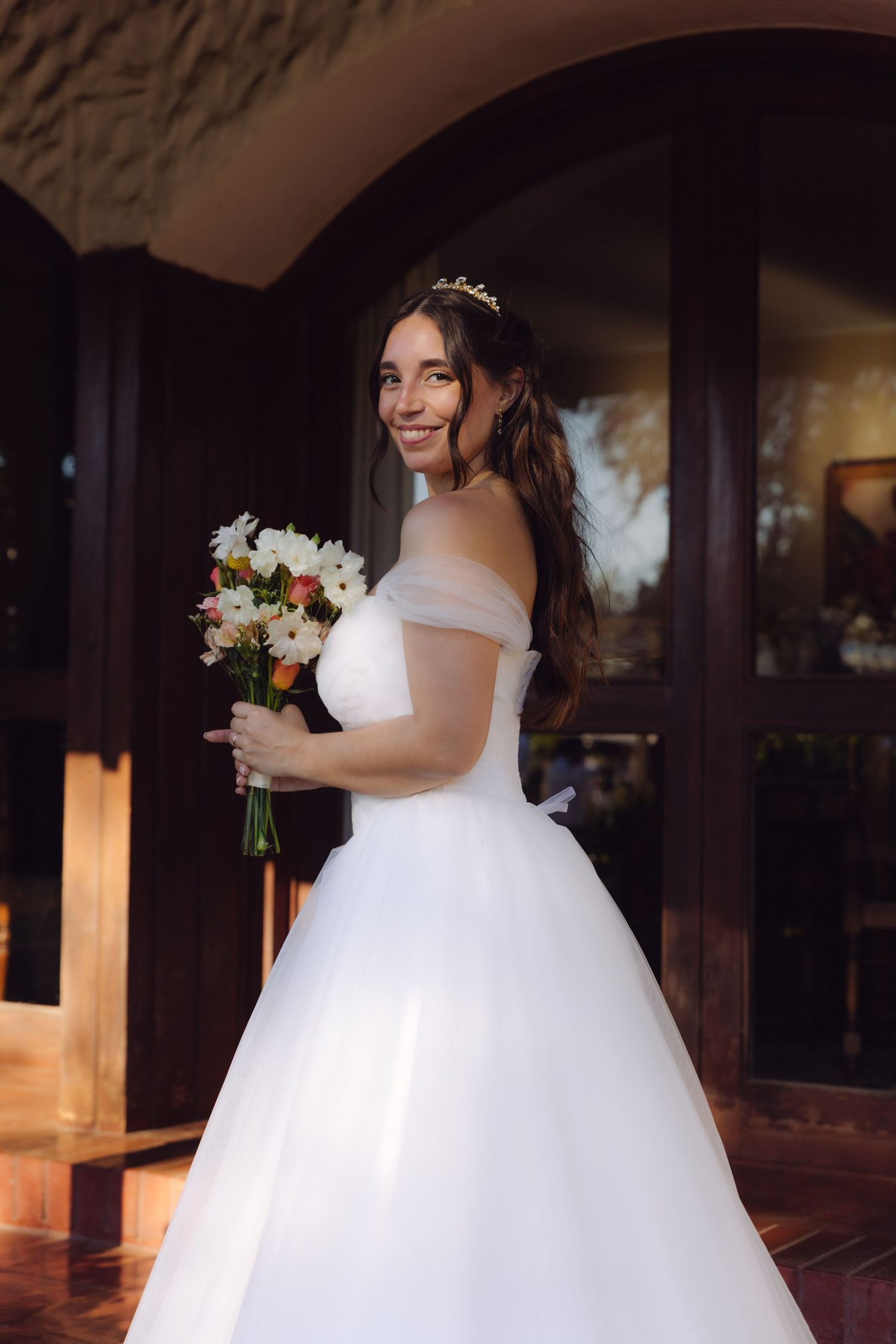 Radiant bride in white ball gown with tiara holds bouquet while posing by arched wooden doors.