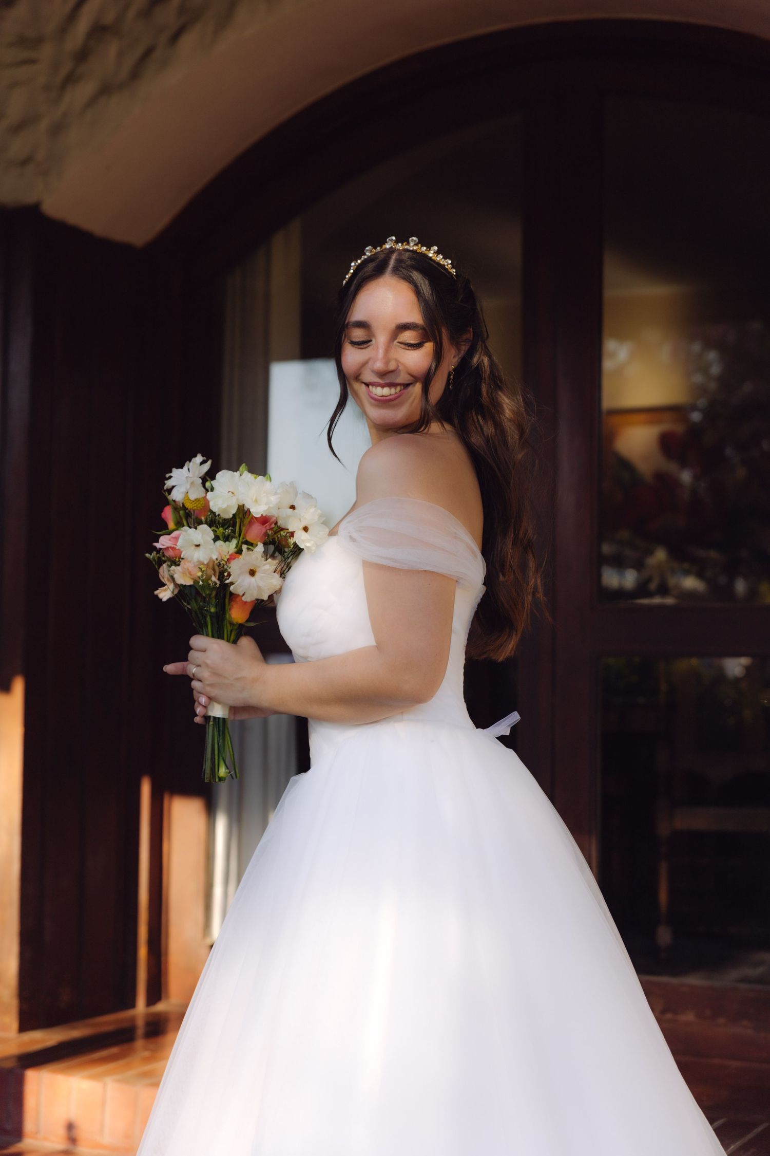 Smiling bride in off-shoulder ball gown holds colorful bouquet near elegant arched doorway.