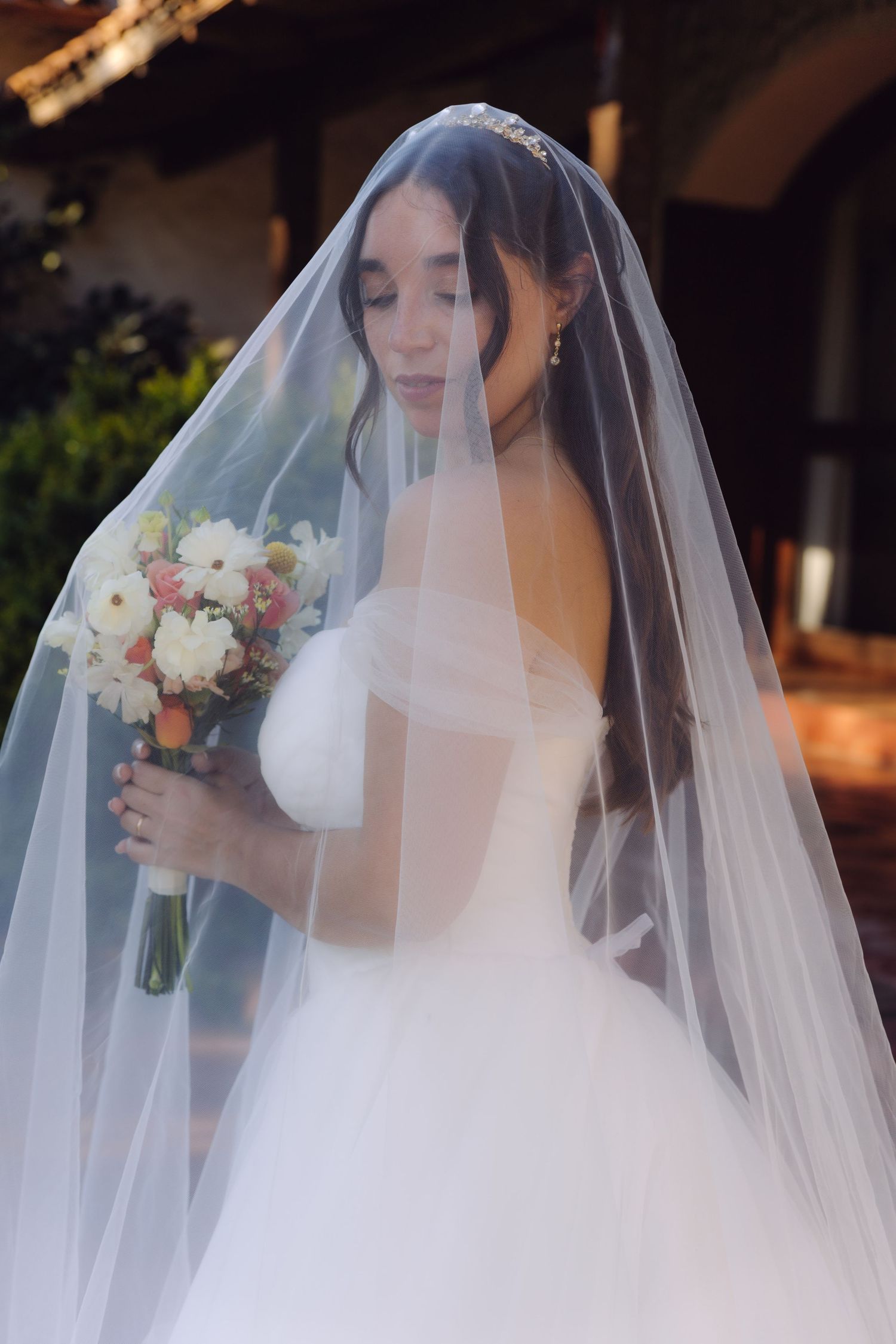 Elegant bride covered by sheer wedding veil holds floral bouquet in soft natural light.