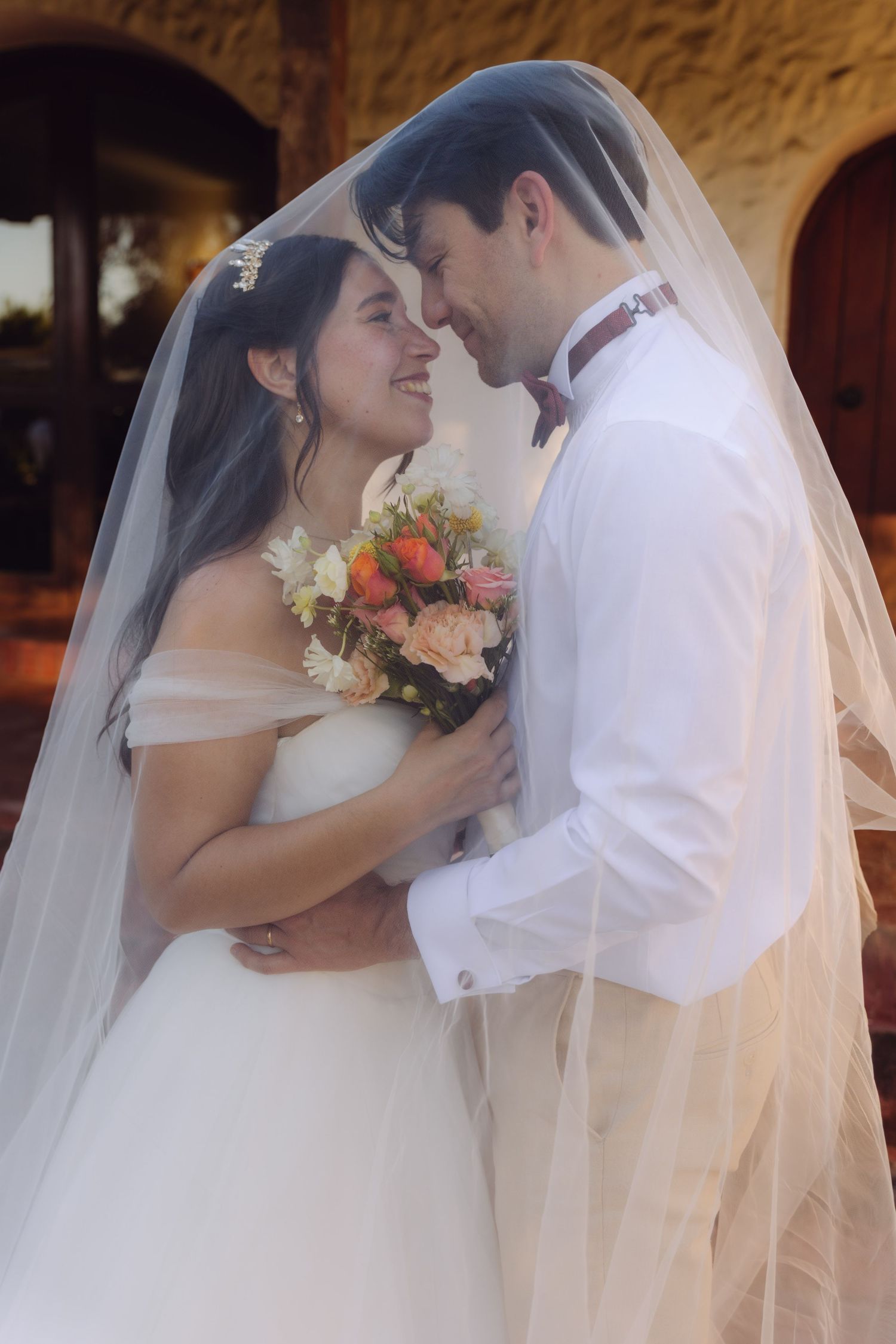 Romantic bride and groom sharing an intimate moment under a wedding veil holding a bouquet.