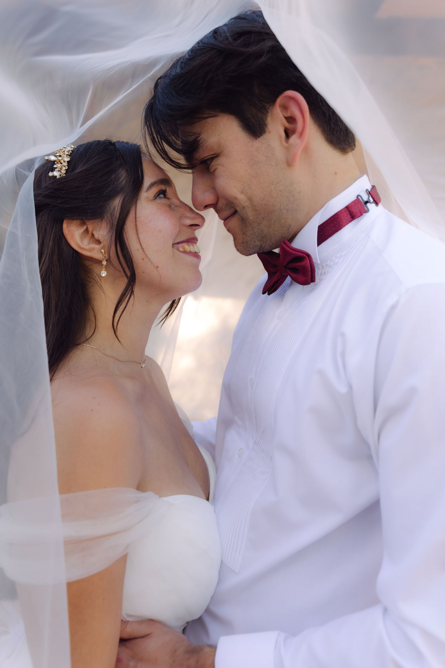 Happy bride and groom share intimate moment under flowing white veil, groom wearing red bow tie.