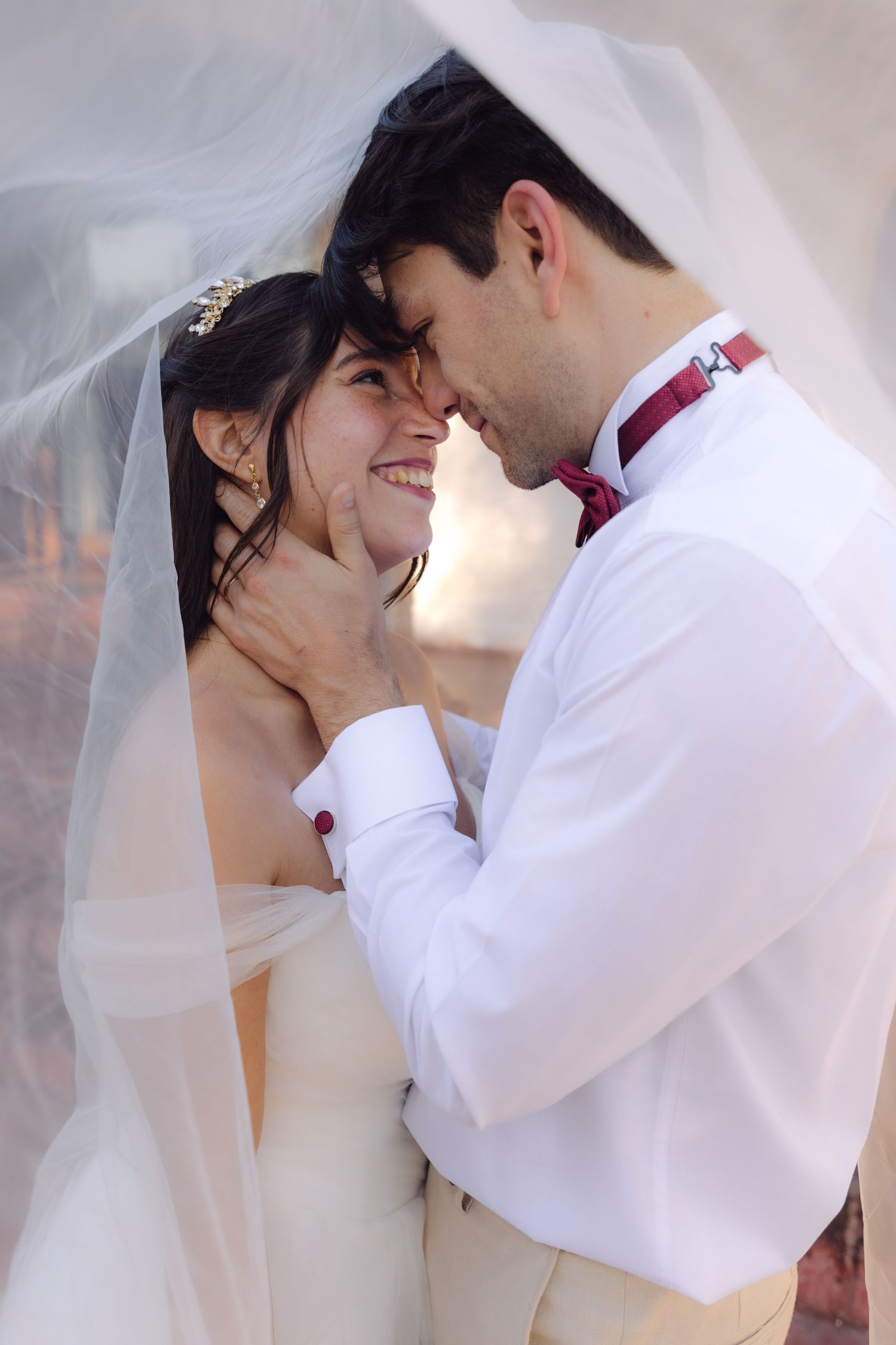 Joyful bride and groom embrace closely under wedding veil, groom in white shirt with red bow tie.