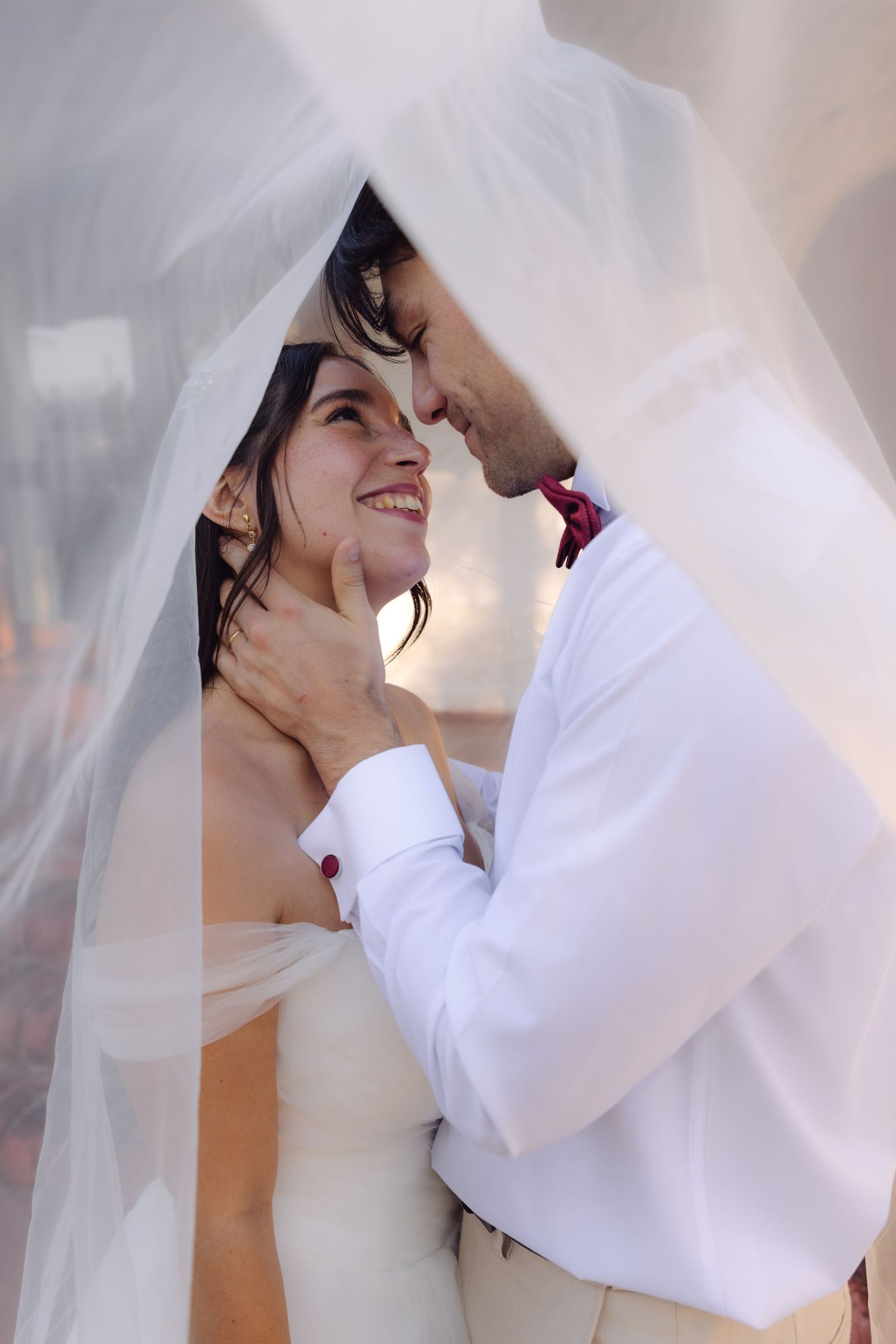 Bride and groom gazing at each other tenderly under a flowing white veil on wedding day.