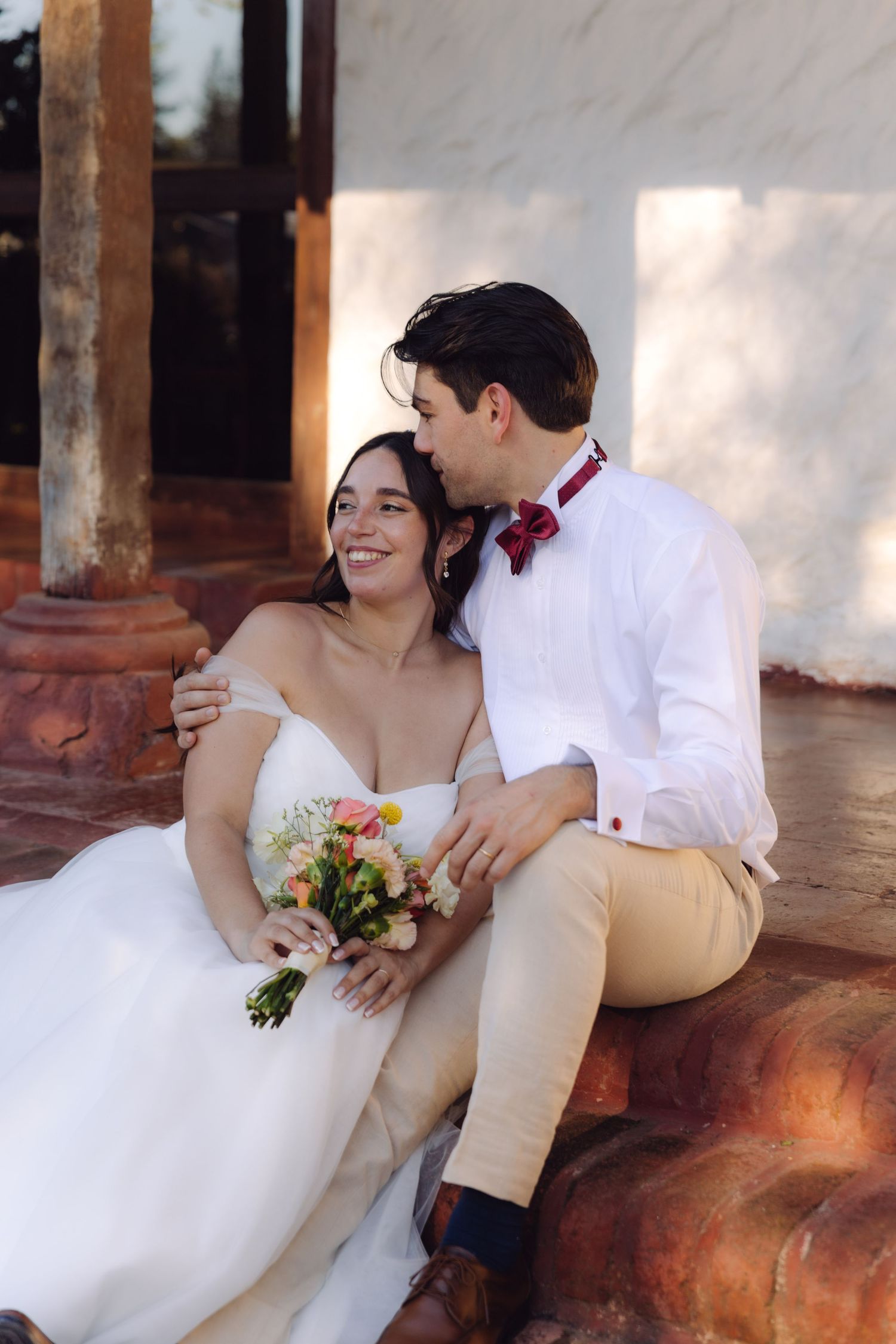 Smiling bride and groom sitting together outdoors, bride holding a pastel floral wedding bouquet.