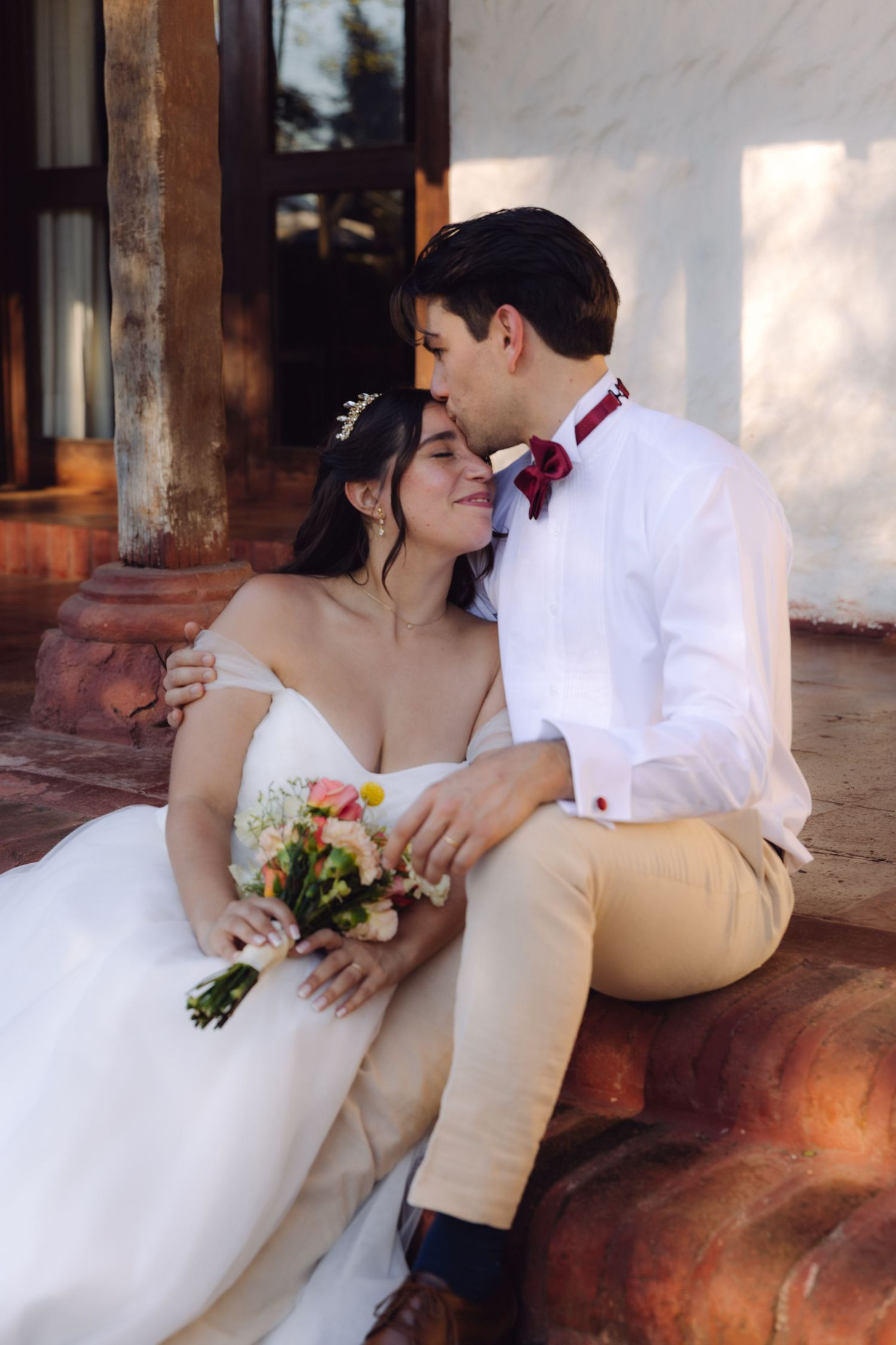Bride and groom sitting on stone steps, sharing an intimate moment, bride holding a floral bouquet in a white wedding dress.