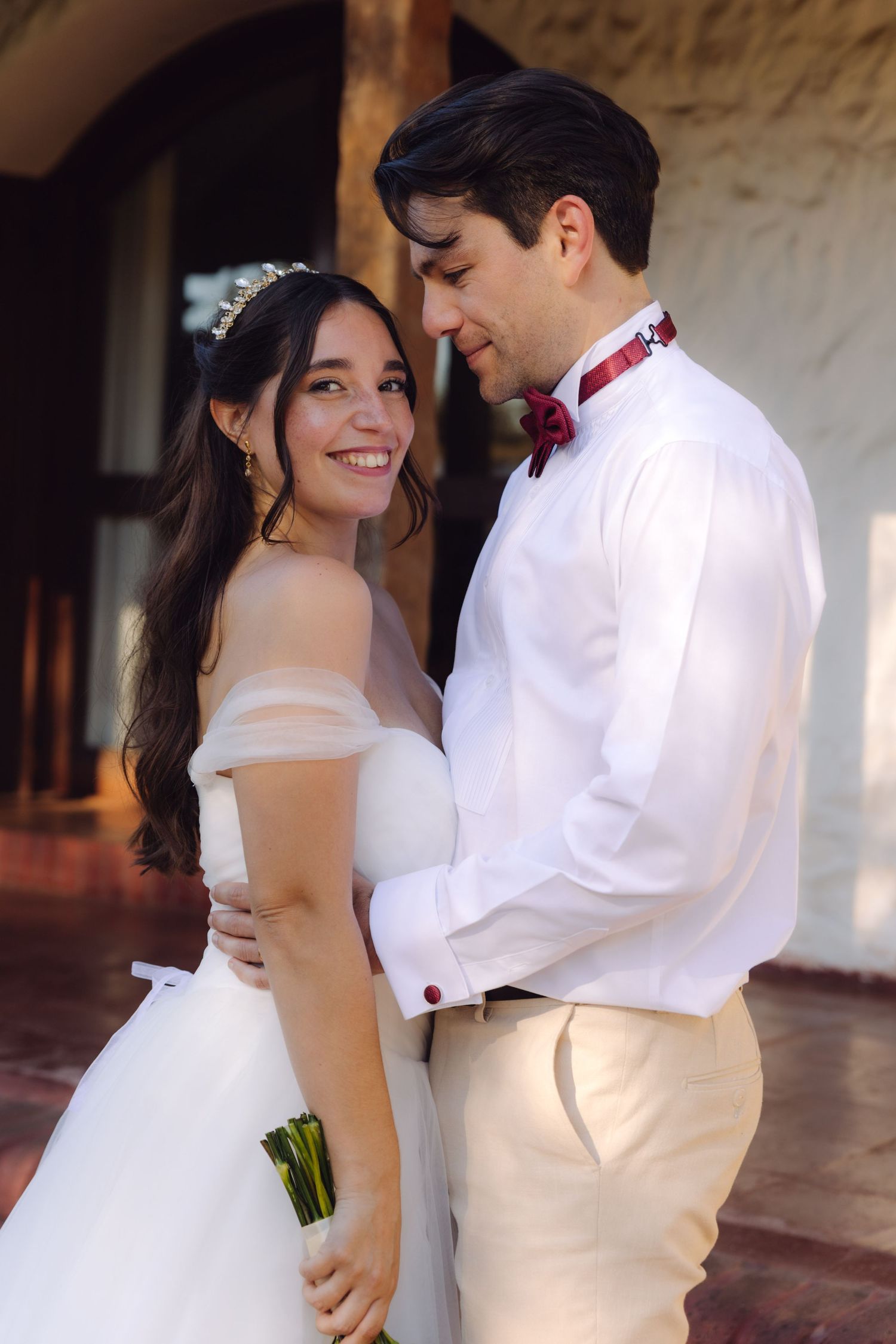 Smiling bride in tiara and off-shoulder gown embraces groom in white shirt with red bow tie.