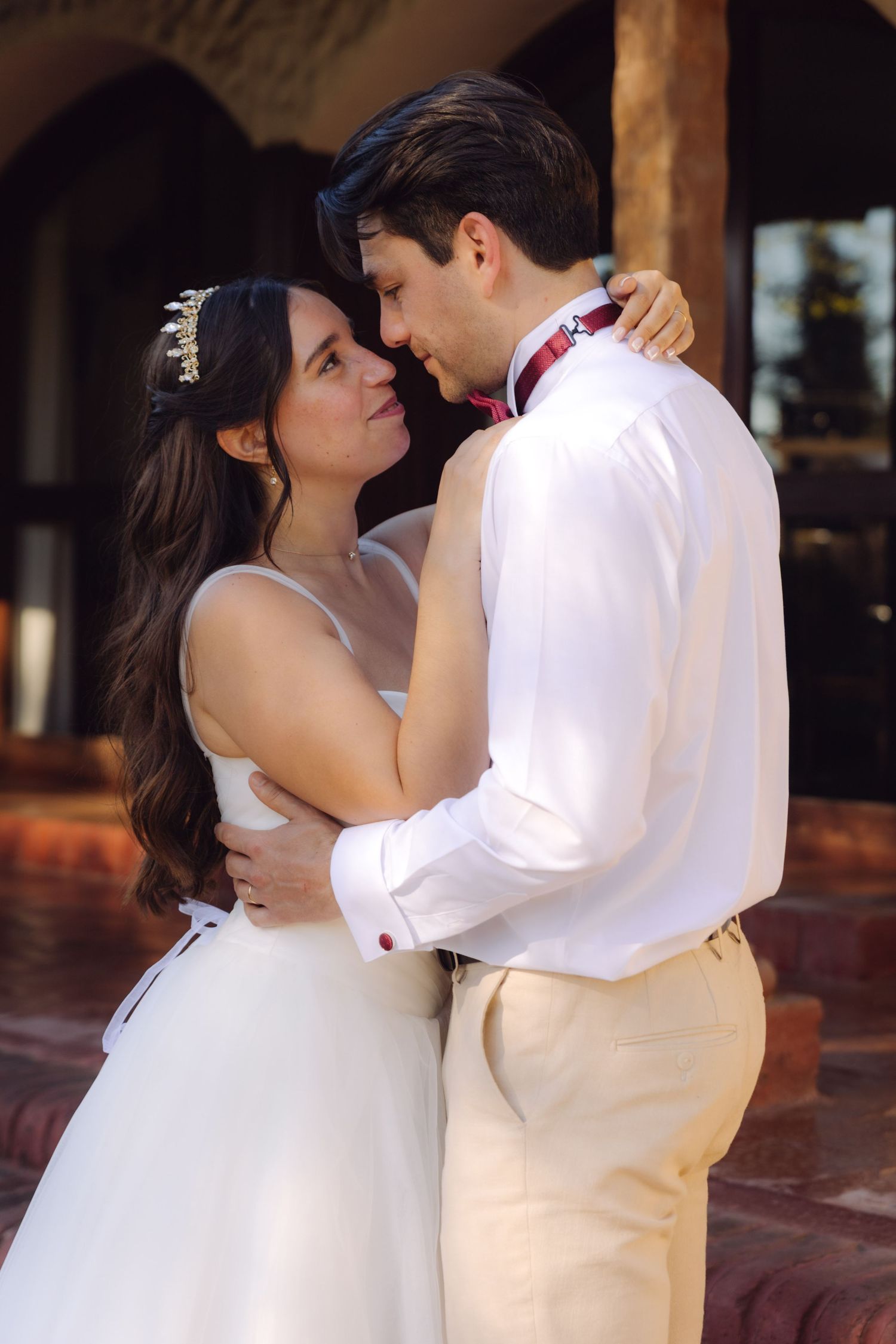 Bride and groom share tender embrace near rustic arched venue, groom in white with red bow tie.