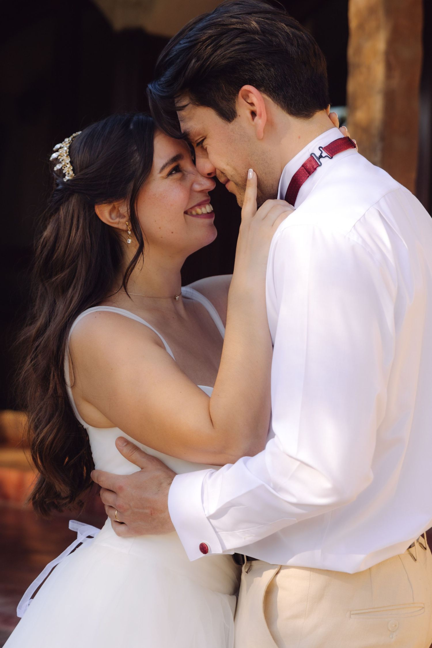 Smiling bride in white dress and groom in white shirt with red bow tie embrace closely outdoors.