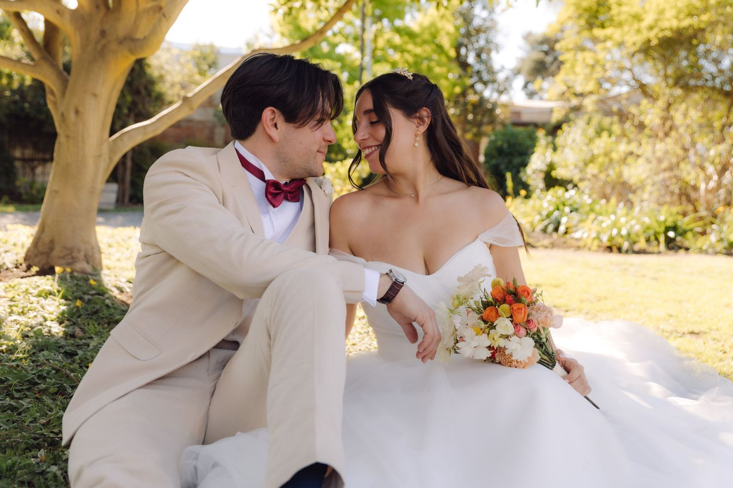 Bride in off-shoulder white gown and groom in beige suit sit together holding colorful bouquet in garden.
