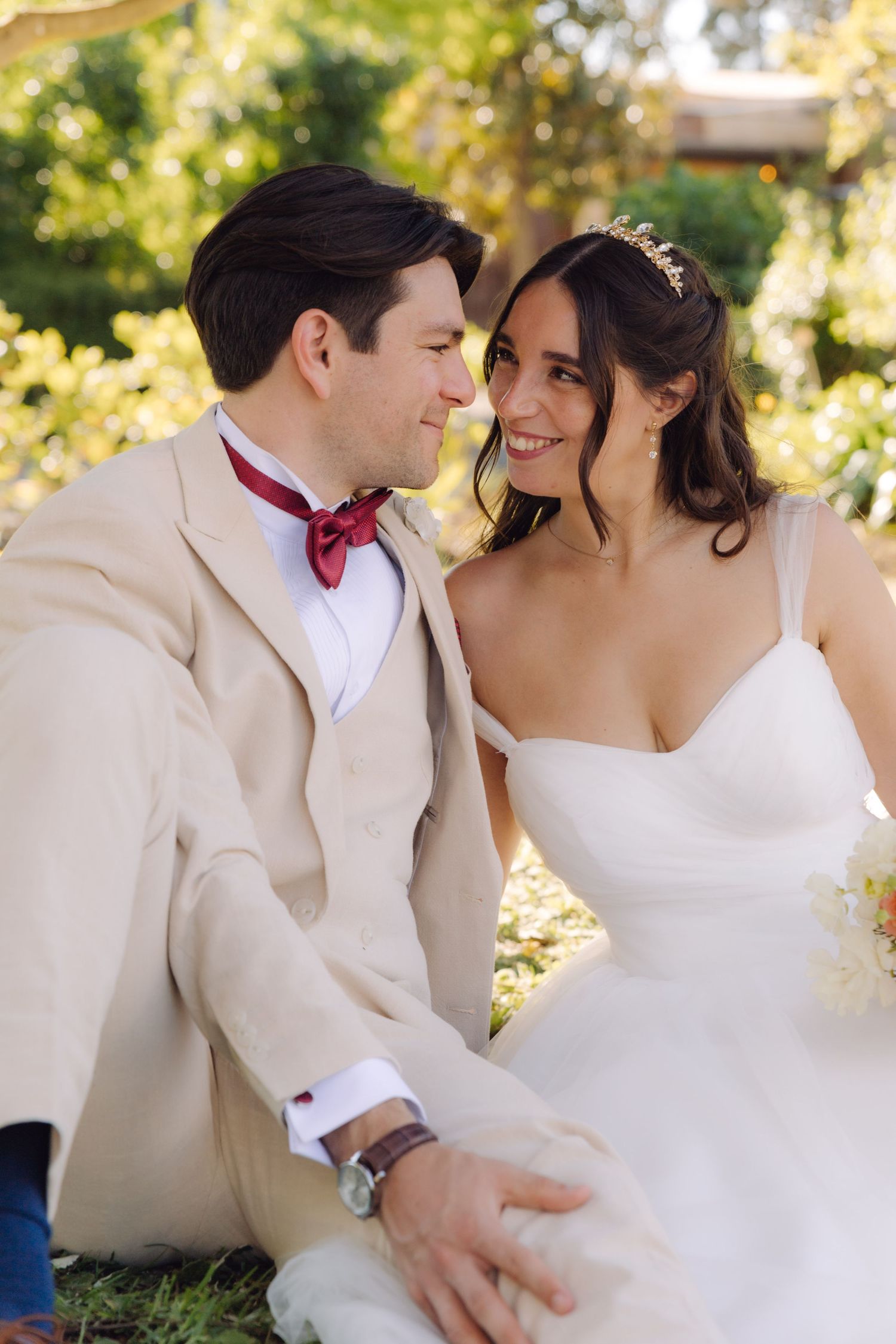 Bride in white dress and groom in beige suit with red bow tie share romantic moment sitting outdoors.