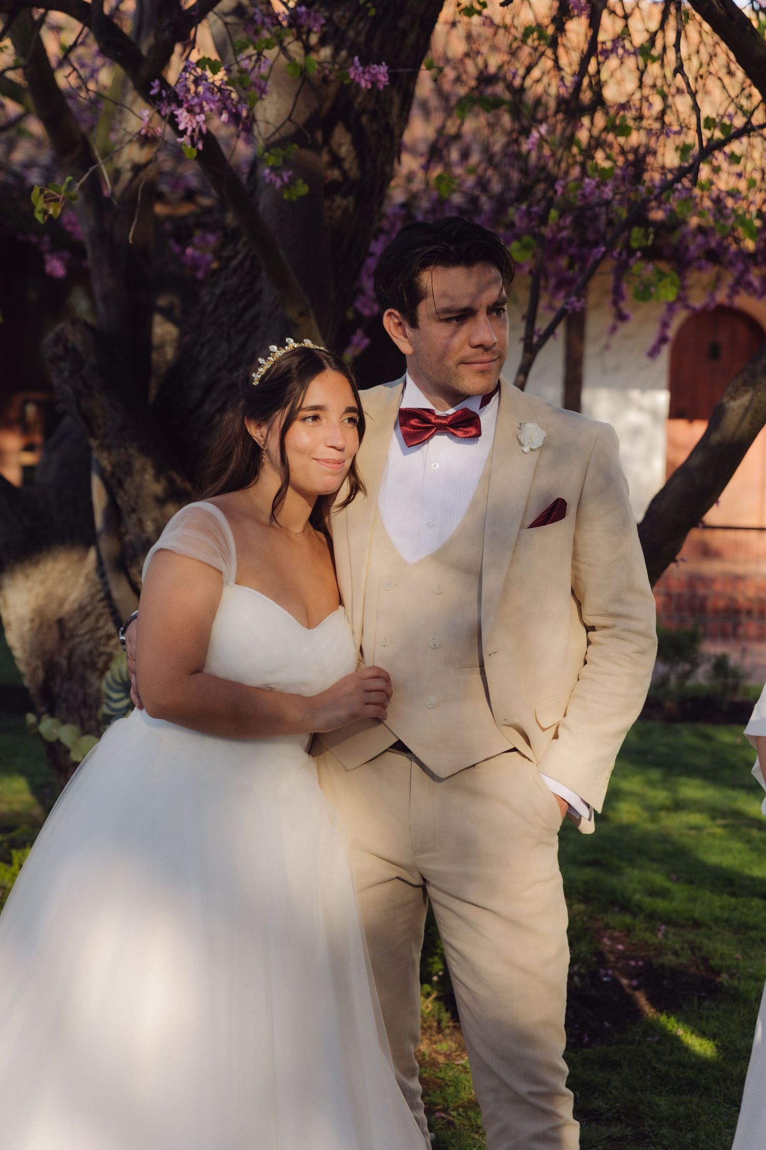 Elegant bride and groom posing together under blooming purple trees, groom wearing a beige suit with red bow tie.