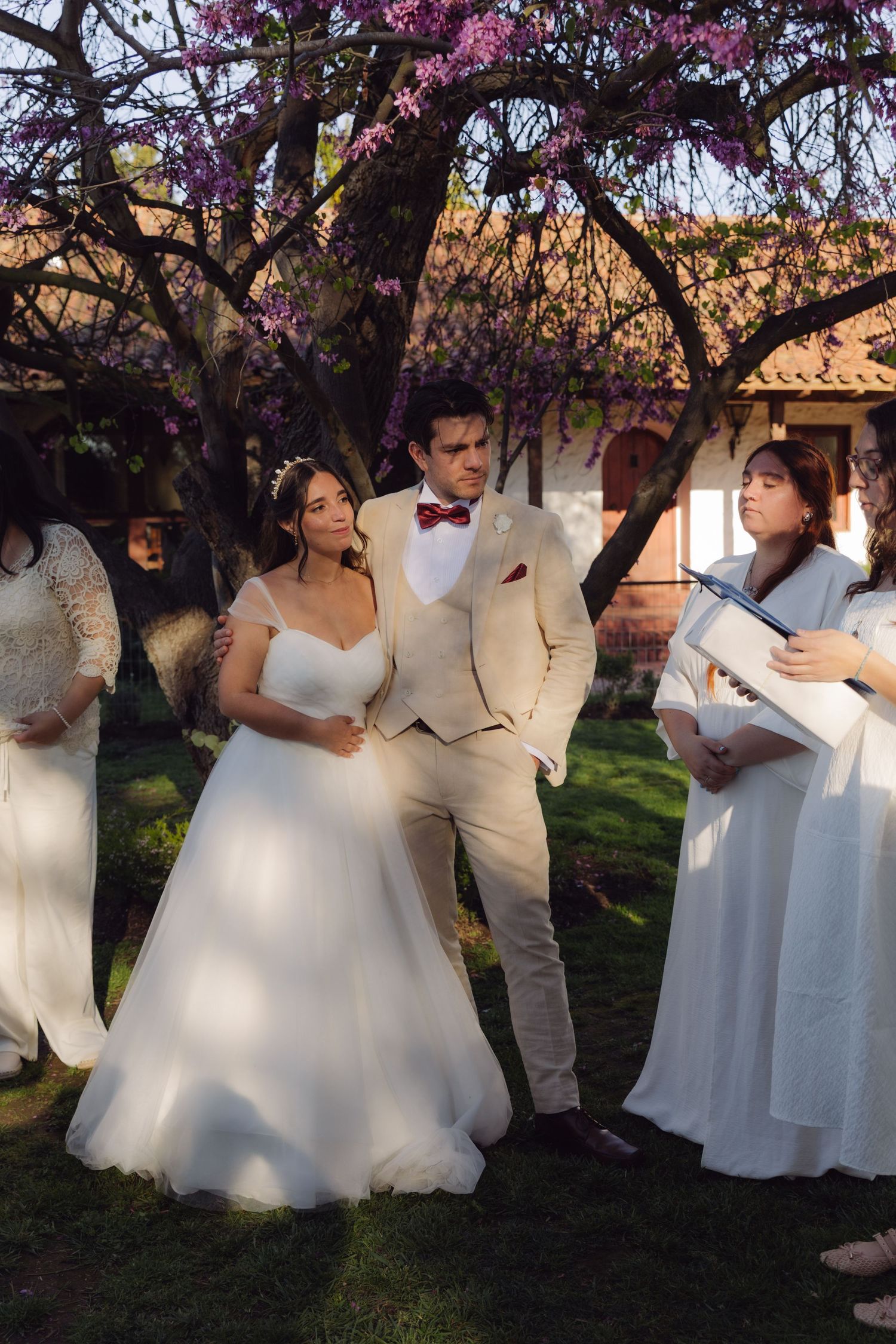 Bride and groom standing during outdoor wedding ceremony under flowering trees with officiant reading from a book.