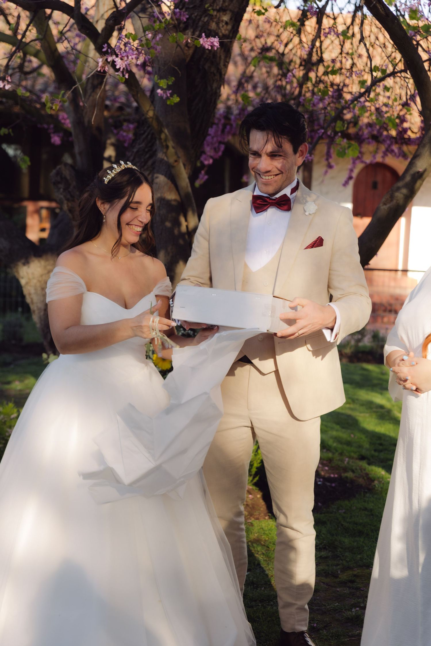 Smiling bride and groom opening a white box together during outdoor wedding ceremony under purple blooming trees.