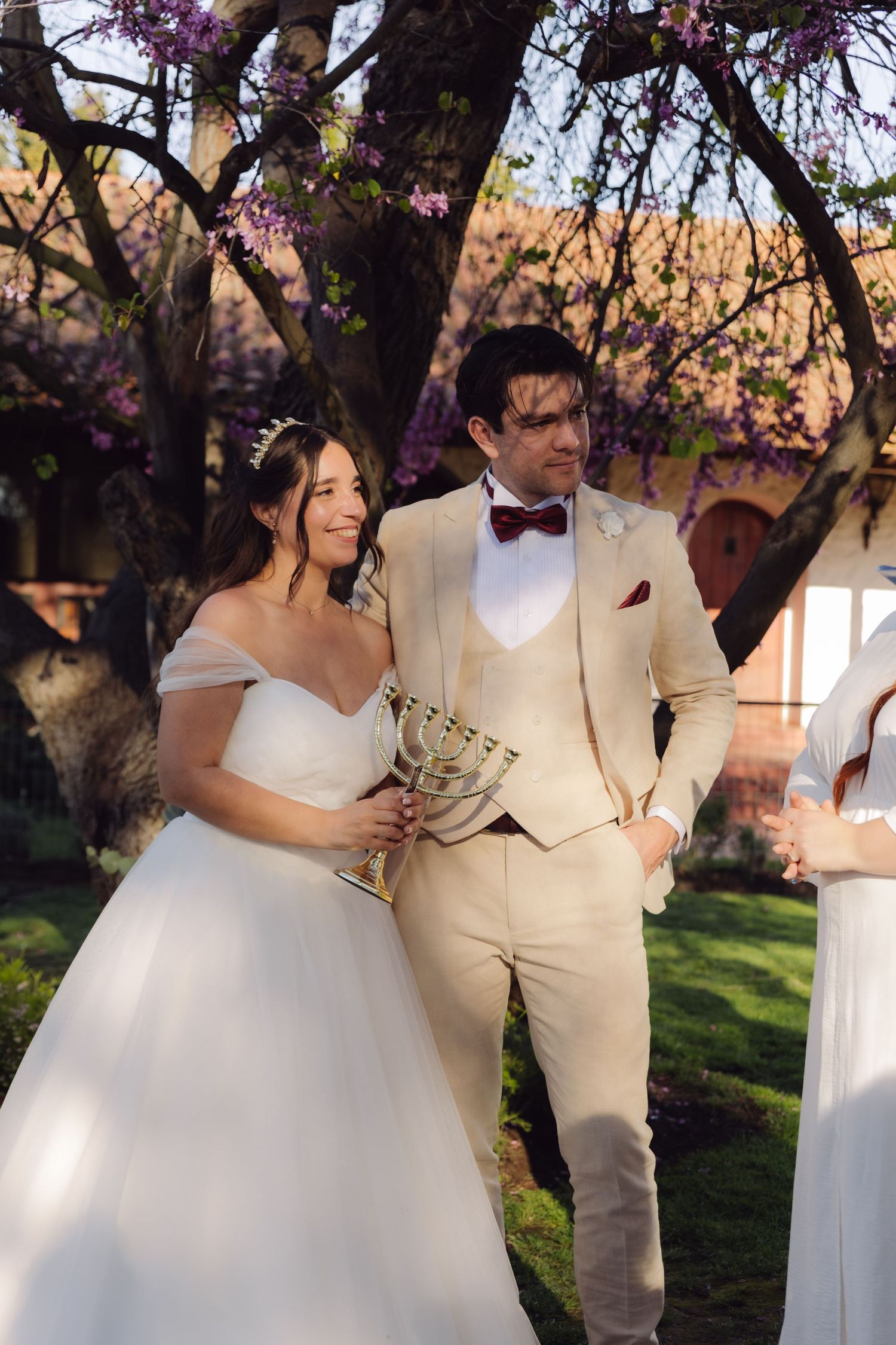 Bride holding a menorah during Jewish wedding ceremony outdoors under blossoming purple trees in golden light.