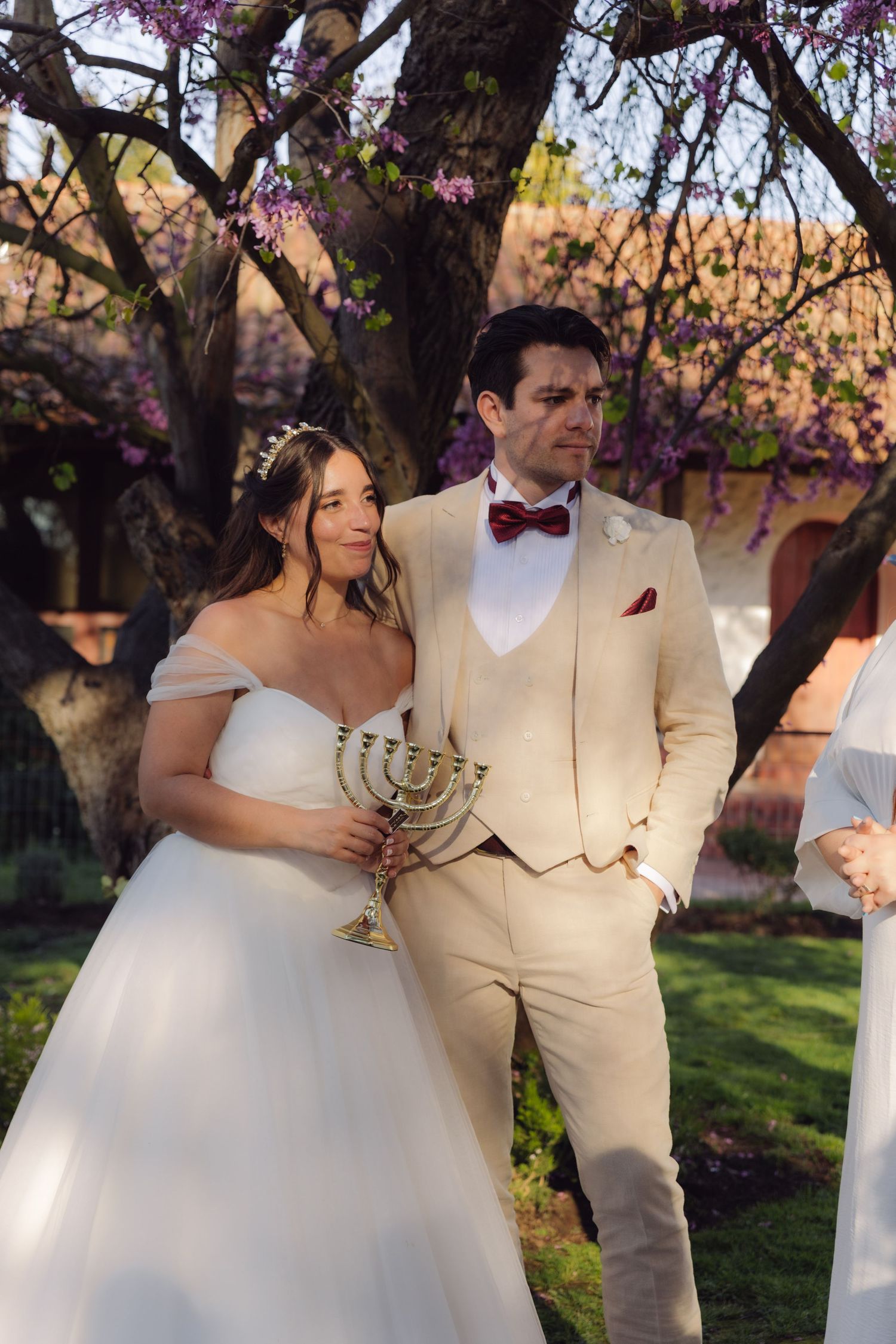 Bride and groom holding a menorah together during outdoor Jewish wedding ceremony beneath flowering spring trees.