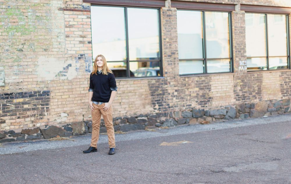 Someone wearing a black sweater and tan shorts stands casually against an industrial brick building exterior.