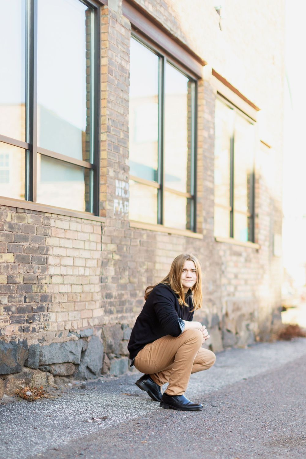 A person in a black jacket and khaki pants crouches near a brick building with large windows.