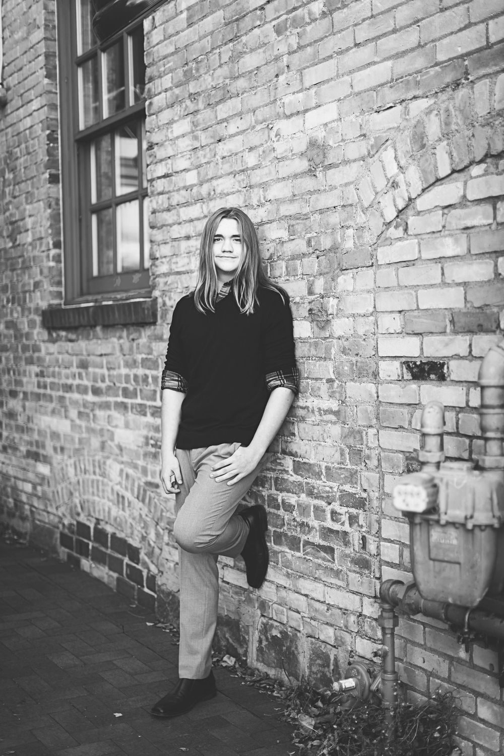 Black and white portrait of a person in a dark sweater leaning against a brick wall in an urban setting.