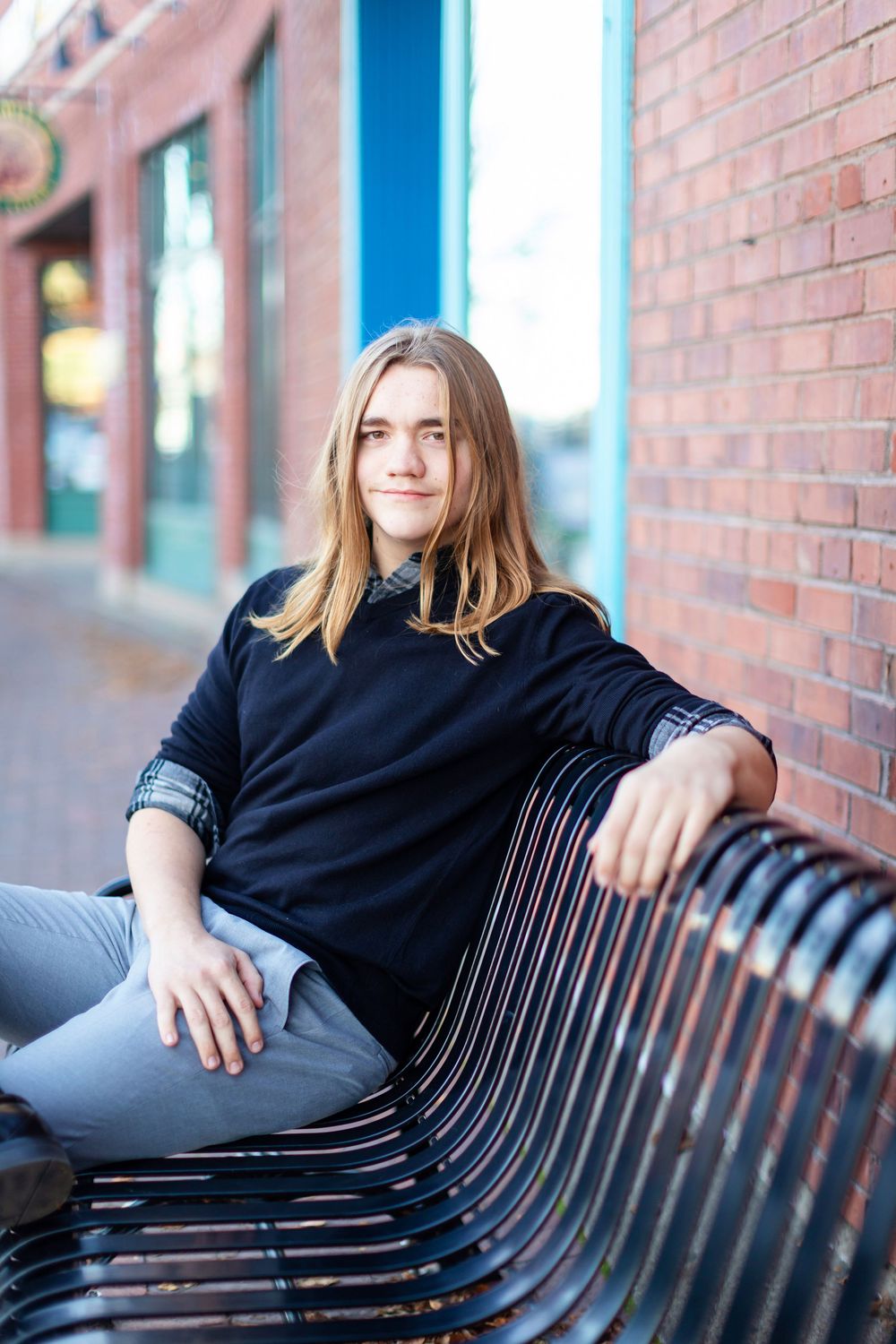 A person in a black sweater sits casually on a metal bench against a brick wall in an urban setting.