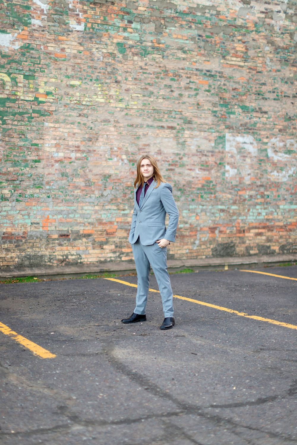 Professional wearing a light gray suit stands confidently against a weathered brick wall in an urban setting.