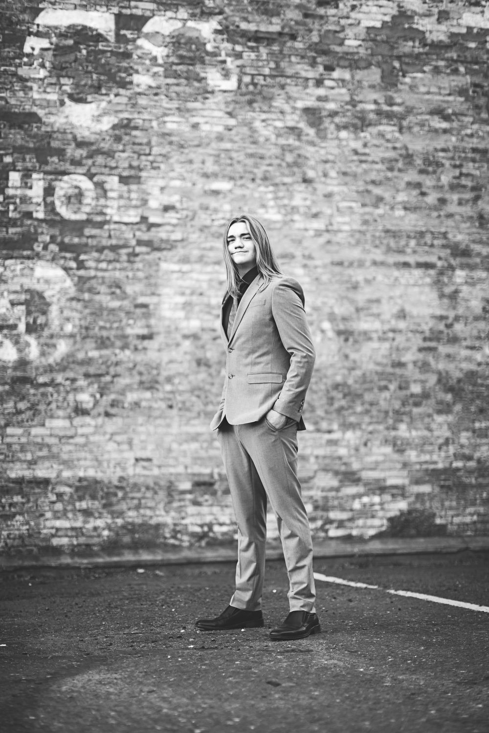 A person in a suit stands confidently against a weathered brick wall in black and white.