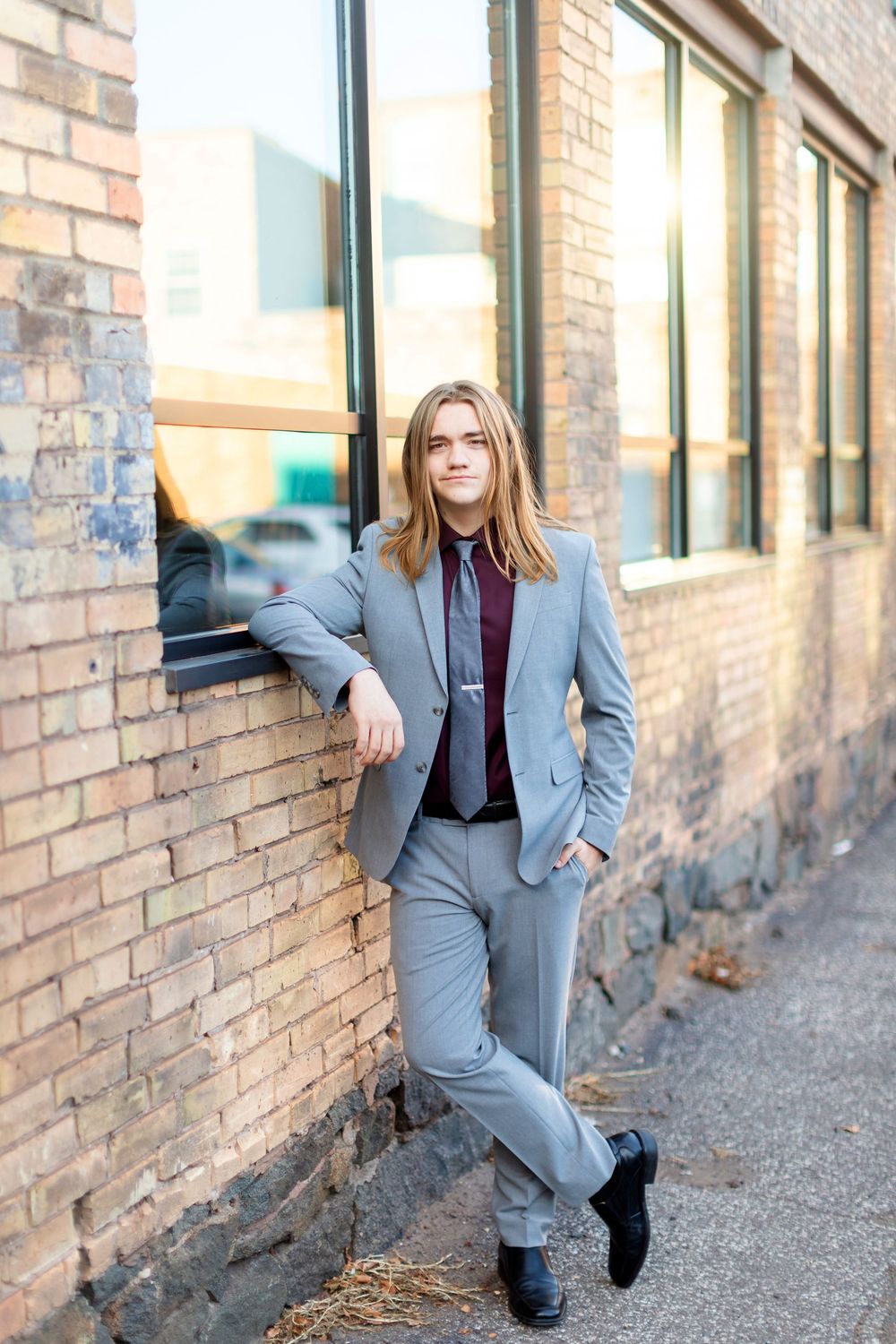 Person in a gray suit and burgundy shirt poses casually against a brick building with sunlit windows.