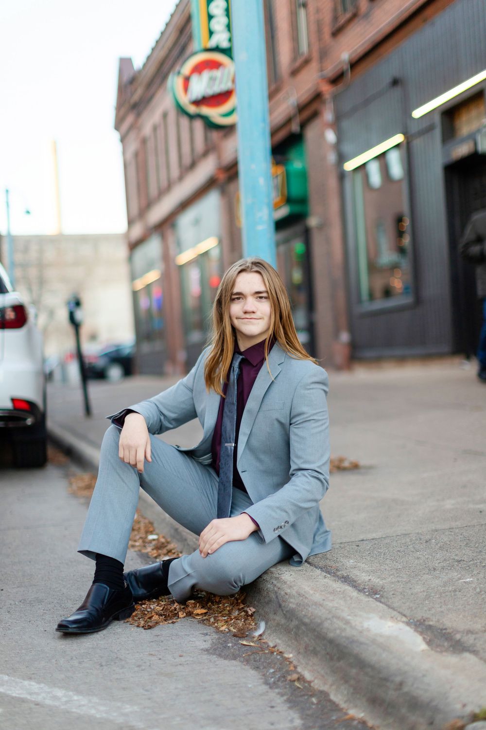 A person in a gray business suit crouches on a city sidewalk near storefronts on an autumn day.